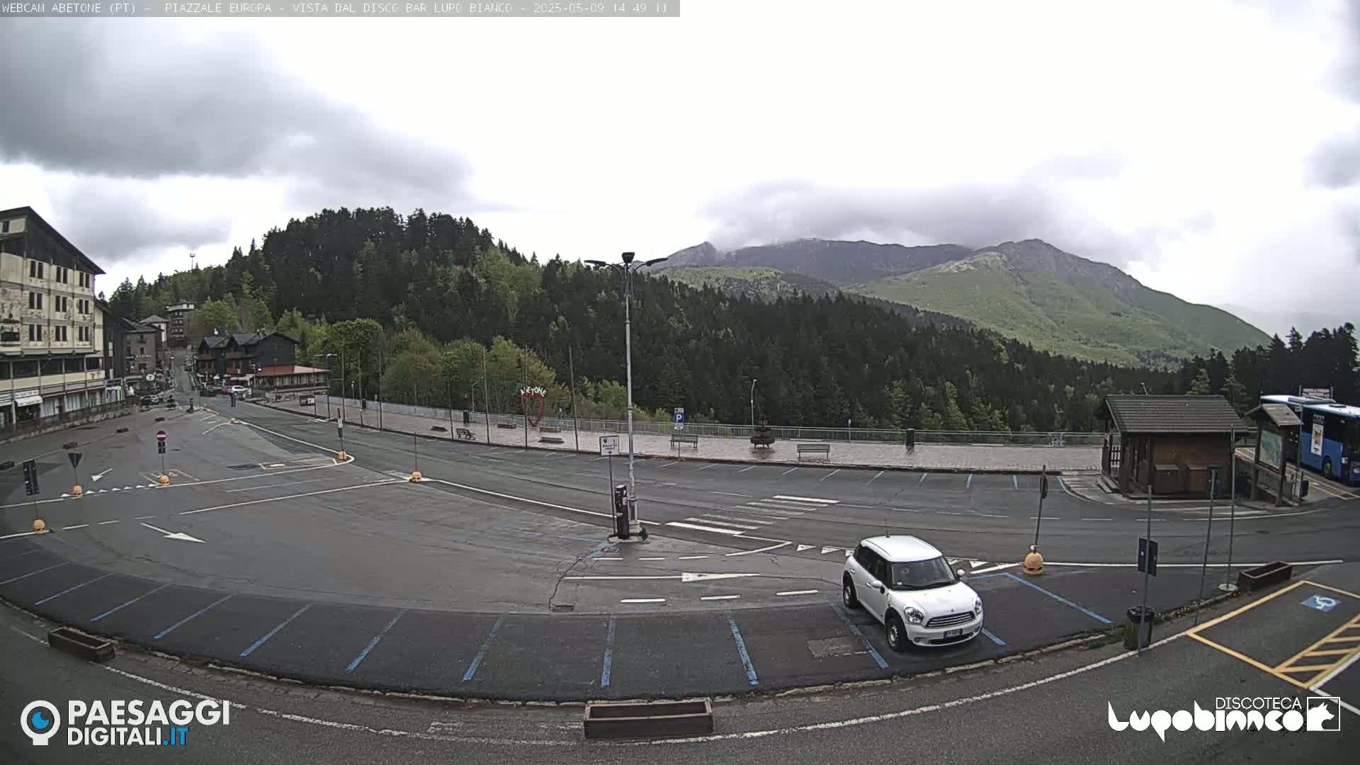 A mostly empty parking lot is situated in a mountainous area under an overcast sky, with a white Mini Cooper parked near a small building and a bus partially visible in the background.