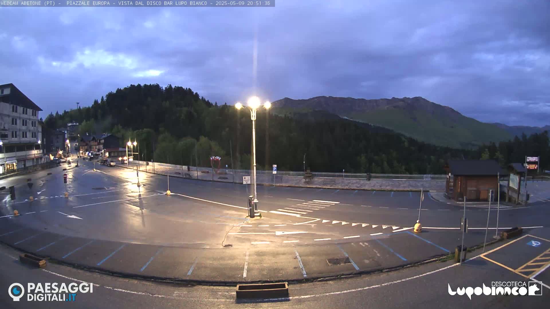 An empty, wet parking lot at dusk is situated in front of a building and overlooks a mountain range under a cloudy sky.