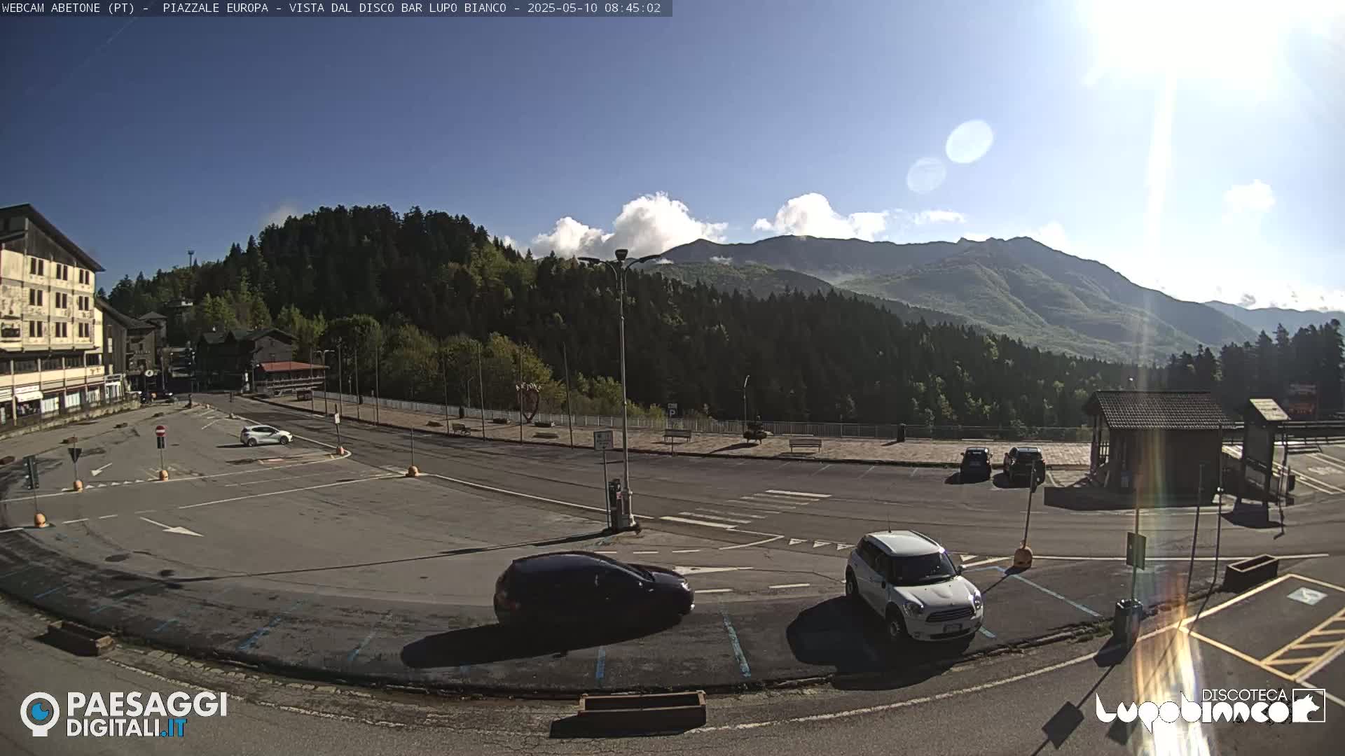 A mostly empty parking lot is situated in front of a building and overlooks a mountain range under a mostly clear sky with a few scattered clouds.
