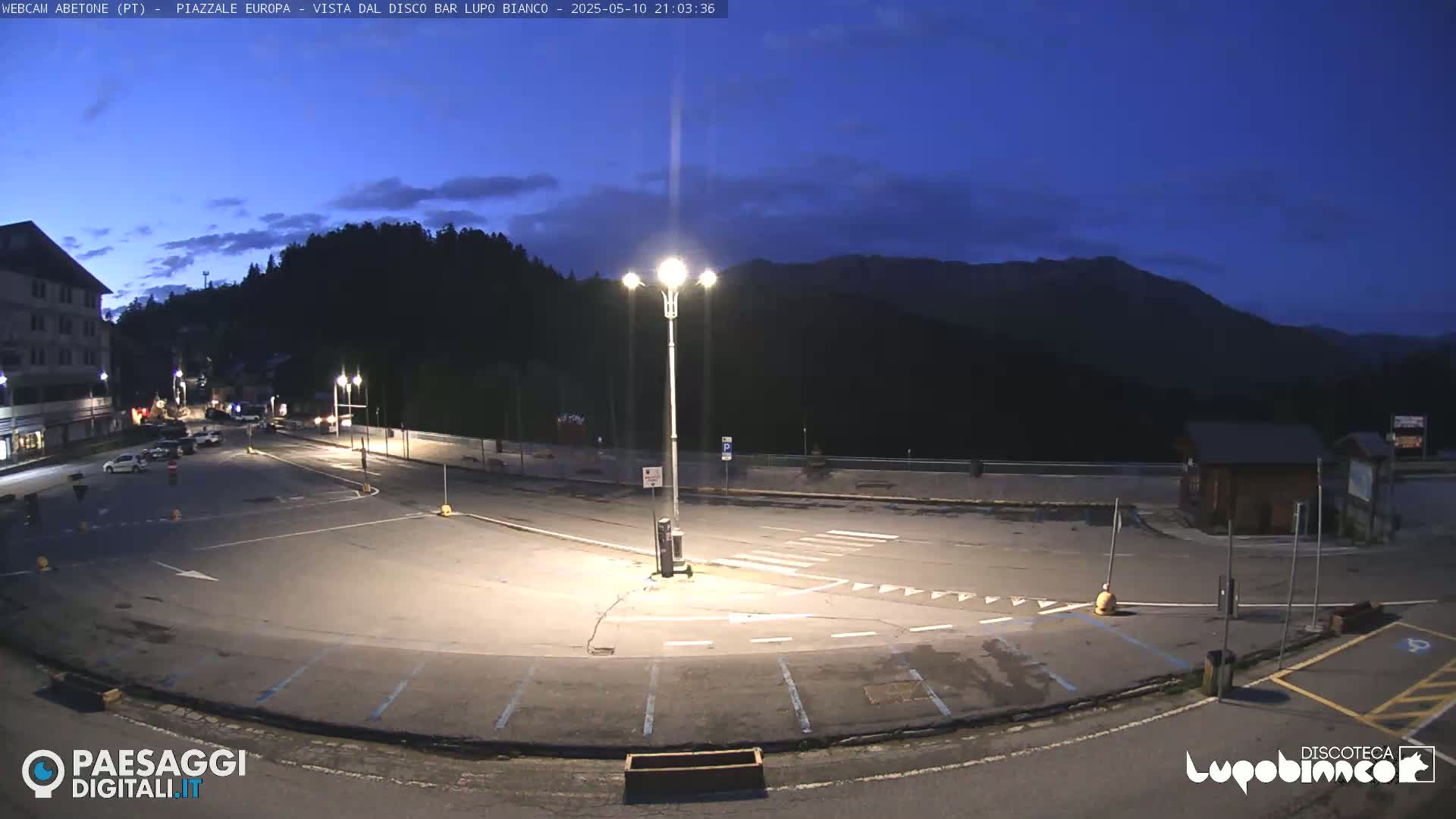 An empty, dimly lit parking lot at twilight, bordered by buildings and mountains under a dark blue sky.