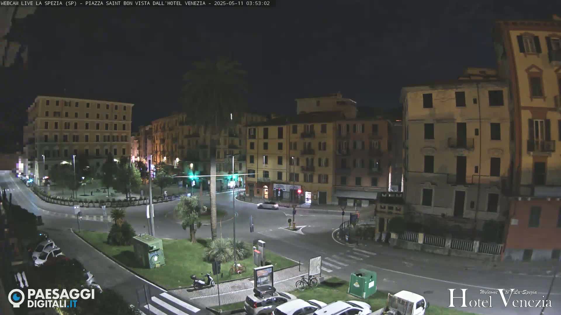 A nighttime view of a city square with several multi-story buildings surrounding it, a few cars parked and driving, and some palm trees.
