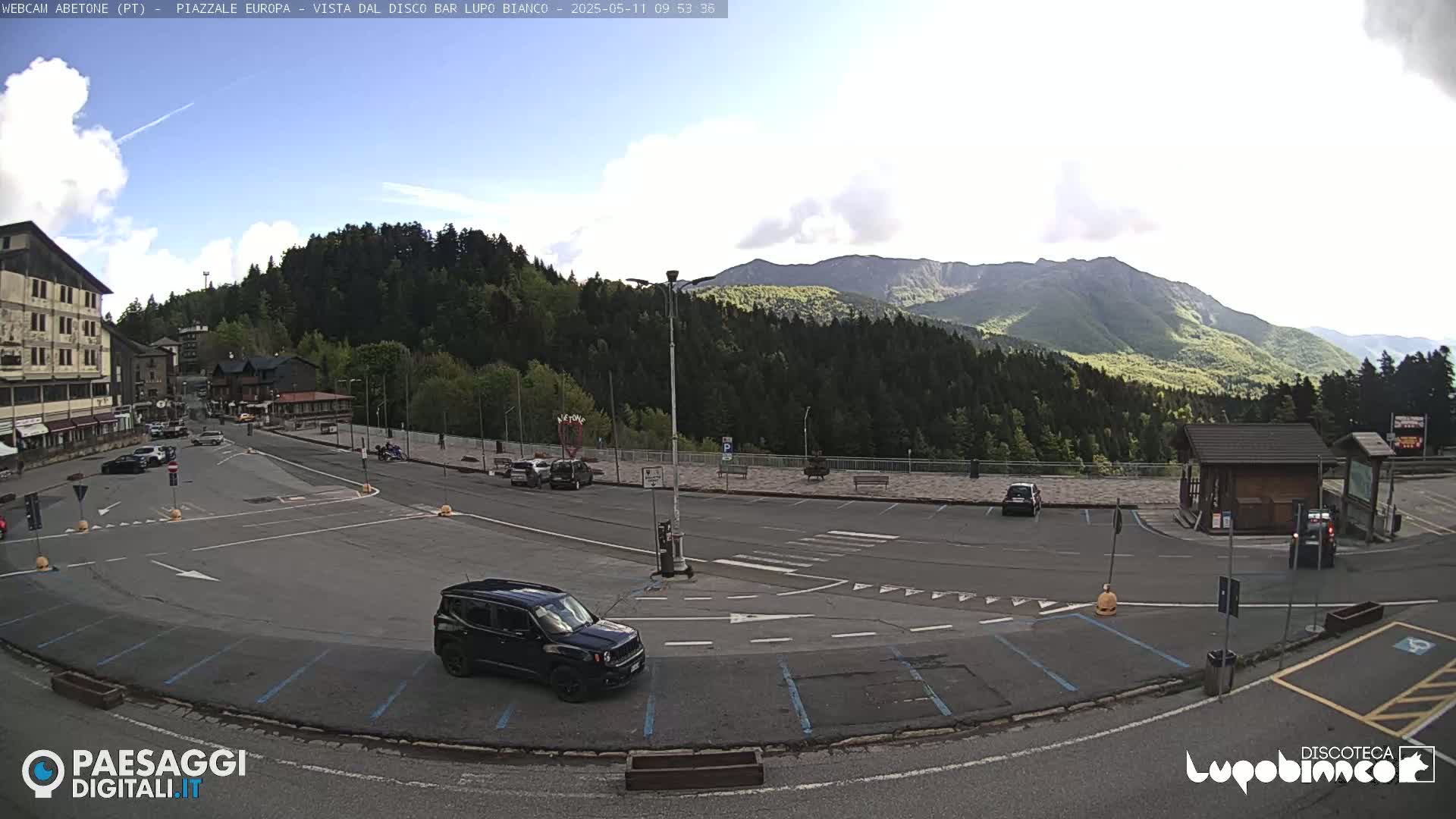 A mostly sunny mountain vista overlooks a mostly empty parking lot and plaza with a few cars and a dark-colored SUV.