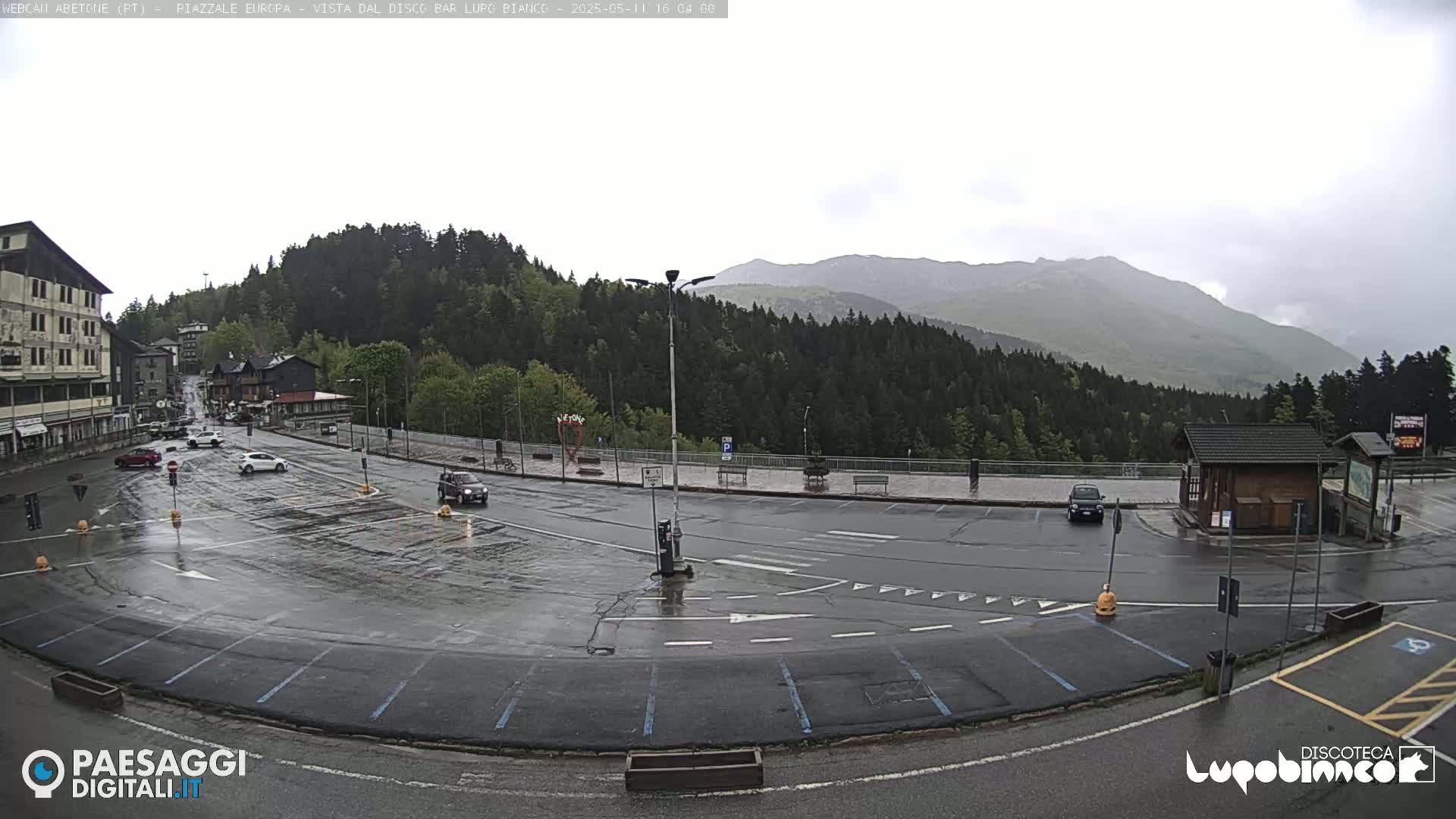 A wet, overcast parking lot and roadway are situated in a mountain valley, surrounded by a dense forest and distant mountains.