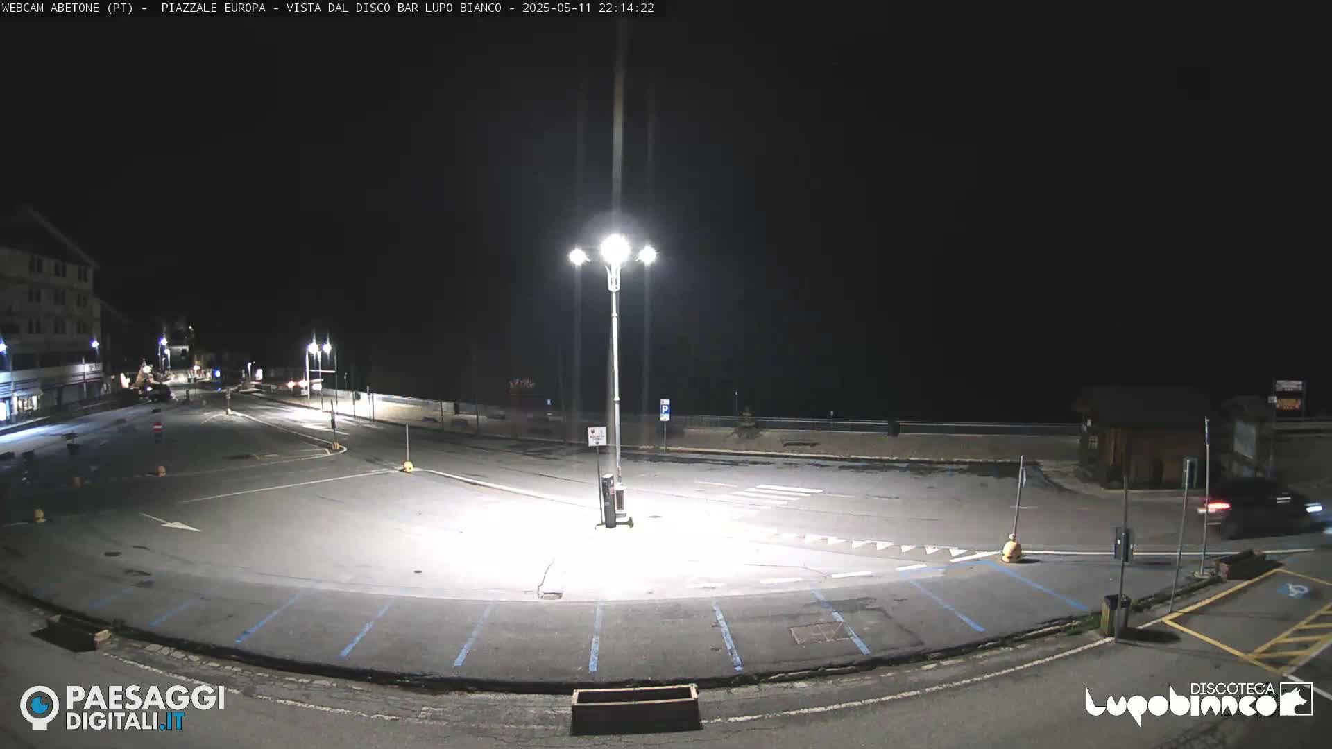 A mostly empty, dark parking lot at night is illuminated by bright streetlights, with a building visible in the background.
