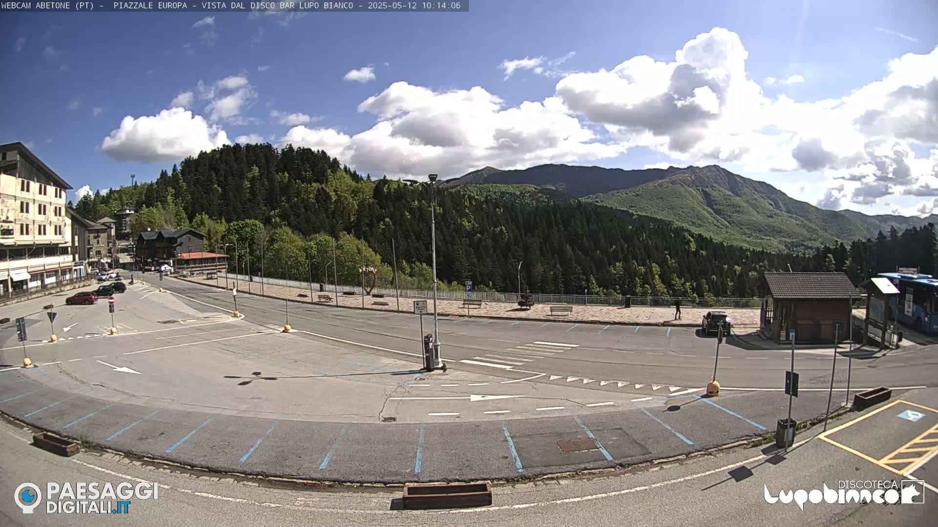 A mostly empty parking lot with light traffic and a few parked cars sits before a mountain range under a partly cloudy sky.