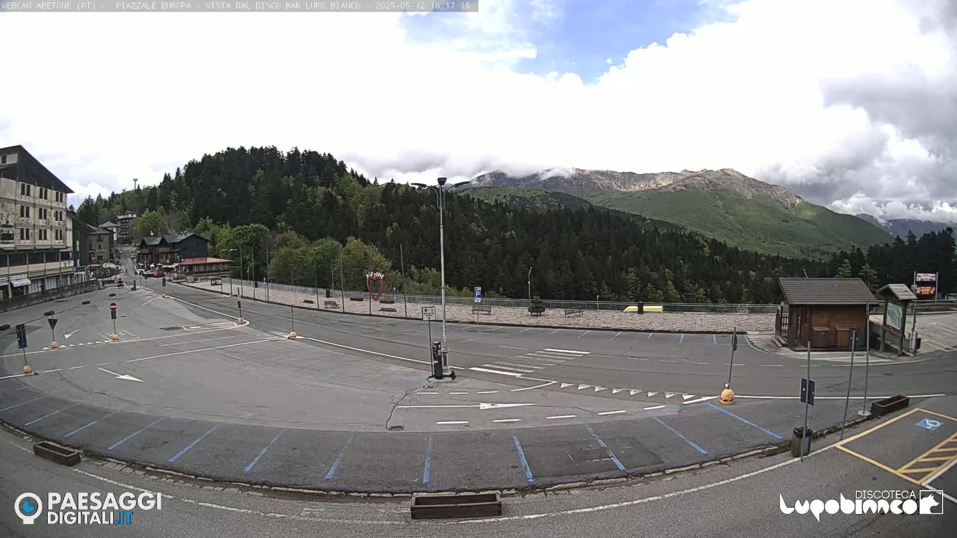 A mostly empty parking lot with light-colored pavement and blue parking lines is situated in front of a building and forested mountains under a partly cloudy sky.