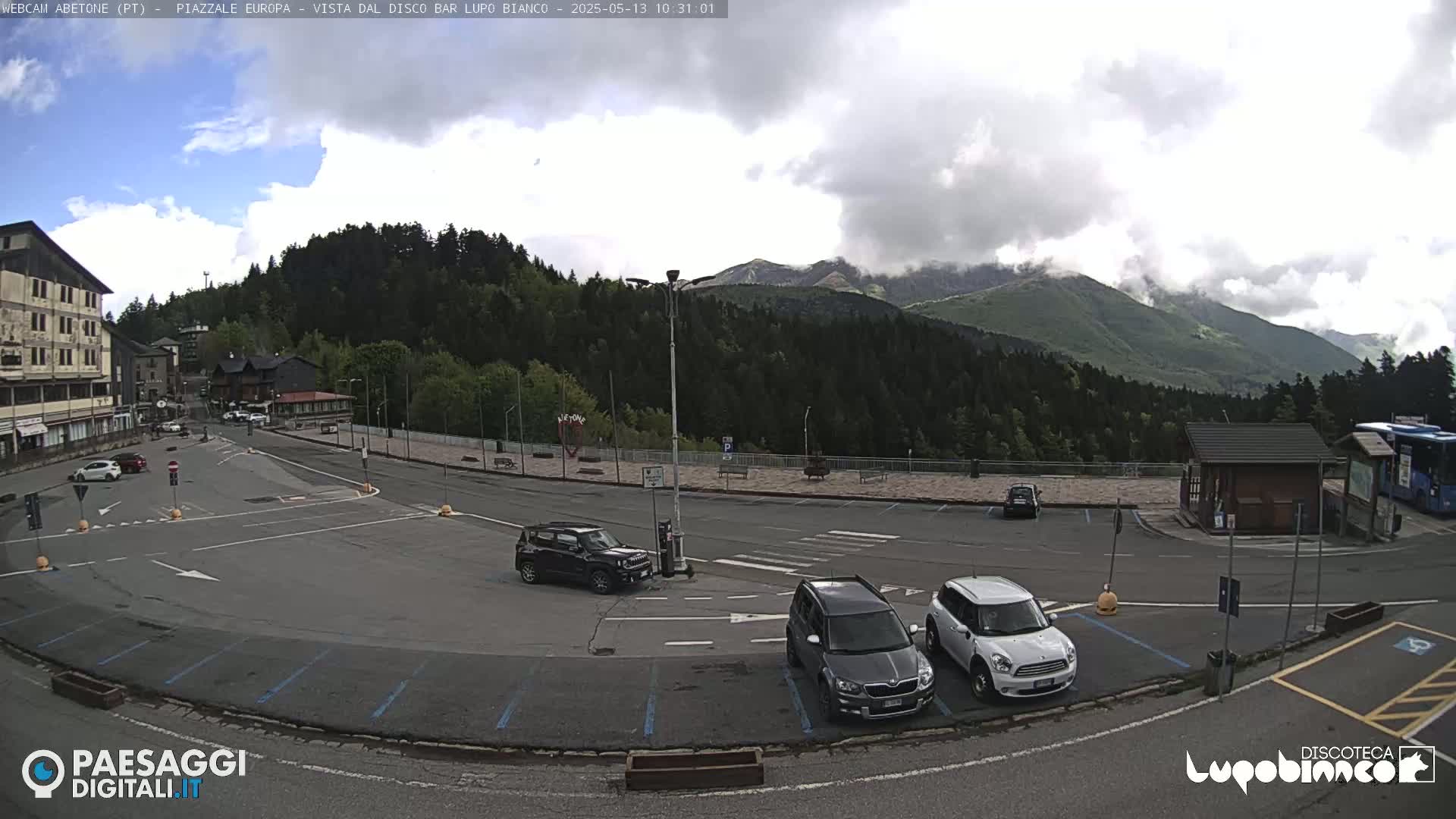 A mostly cloudy day shows a mountain view overlooking a mostly empty parking lot with several cars, a small building, and a few buildings in the distance.
