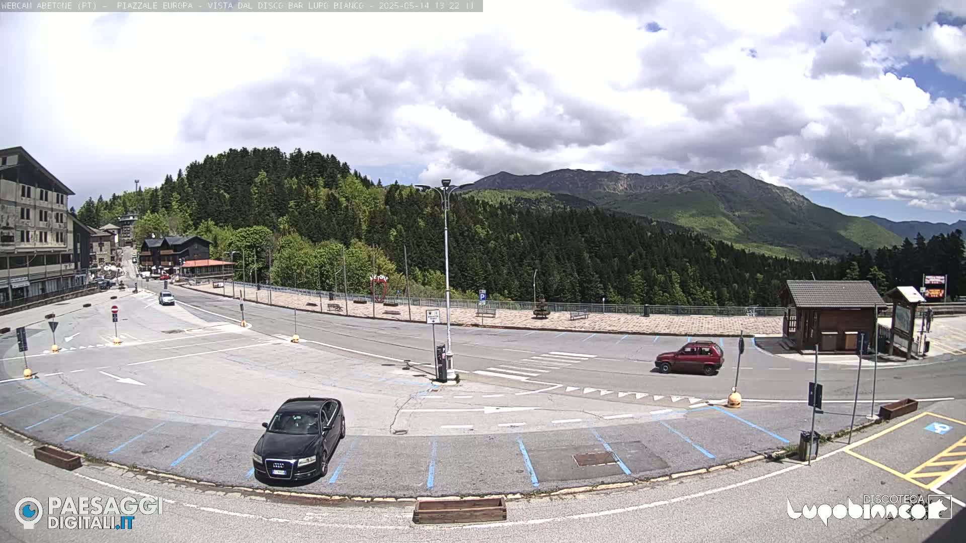 A mostly sunny parking lot with a few cars, overlooks a mountainous landscape covered in lush green forests.