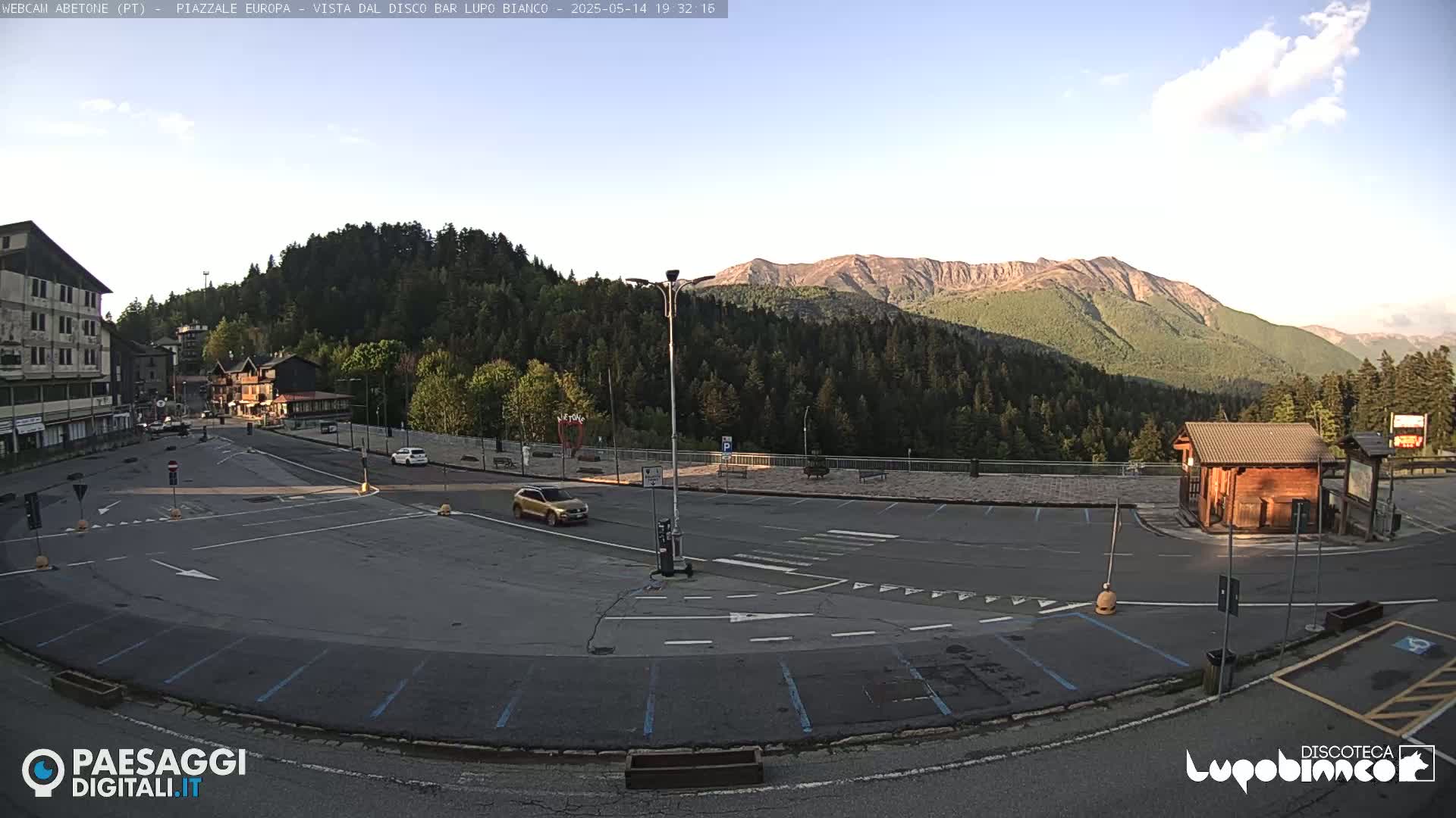 An empty parking lot with a few cars and buildings is situated in front of a mountain range under a clear, bright sky.