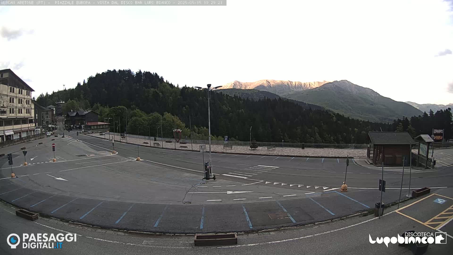 An empty parking lot with light-colored pavement and blue parking space lines is situated in front of a large building and a mountain range under a mostly clear, light sky.