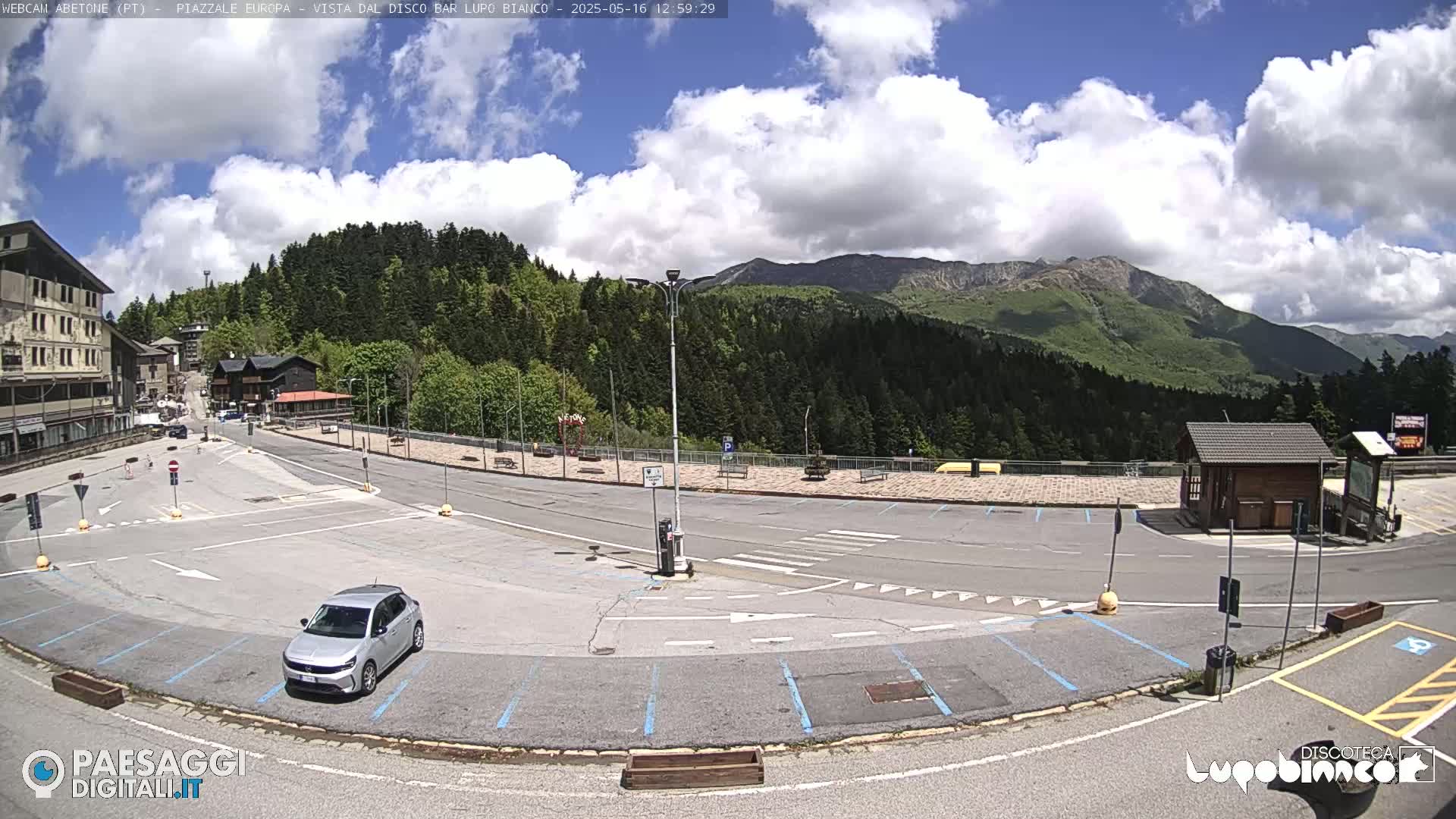 A mostly empty parking lot with a silver car, situated in front of a mountain range under a partly cloudy sky.