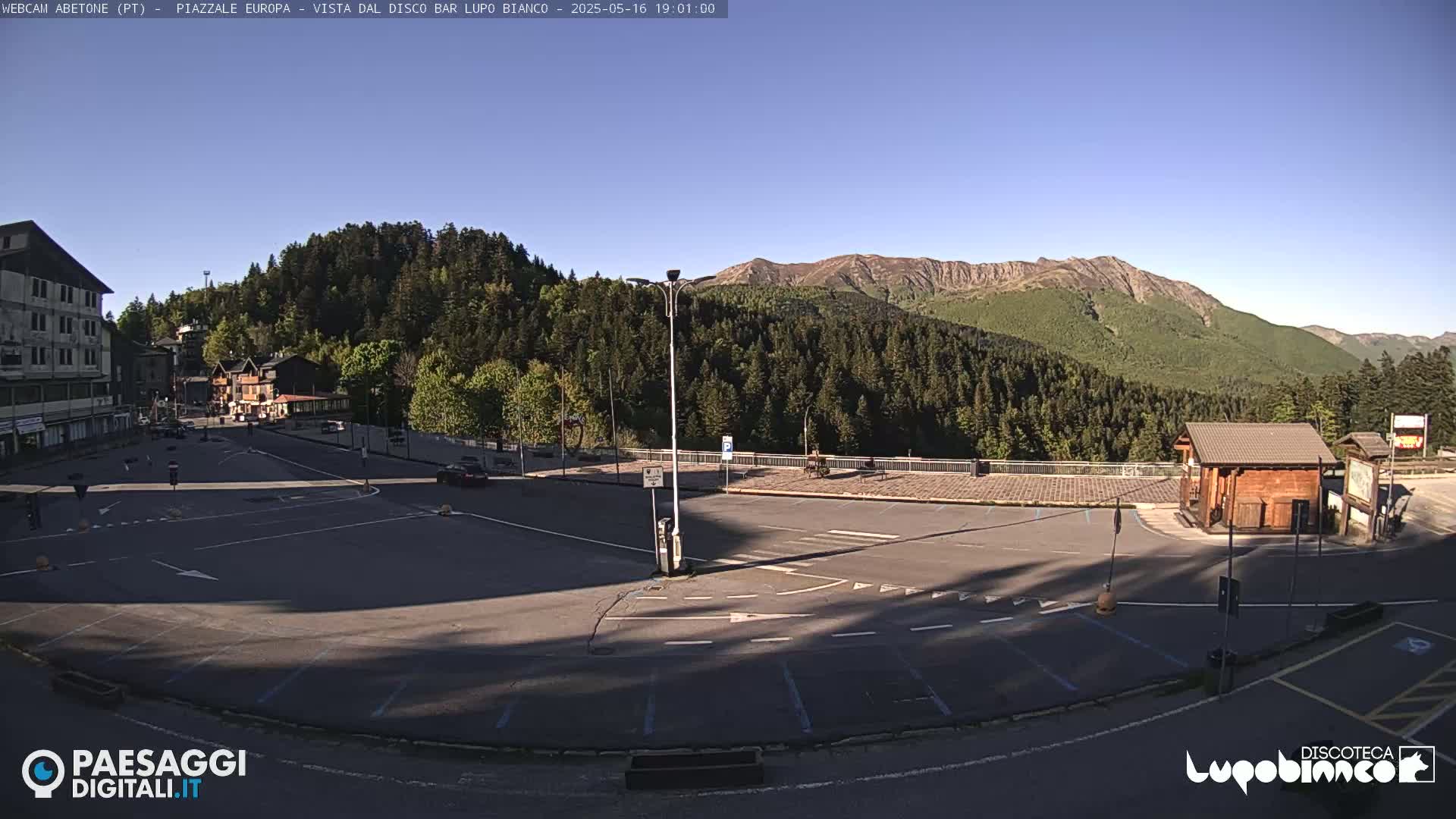 An empty parking lot overlooks a mountain range under a clear, sunny sky;  a small building is visible to the right.