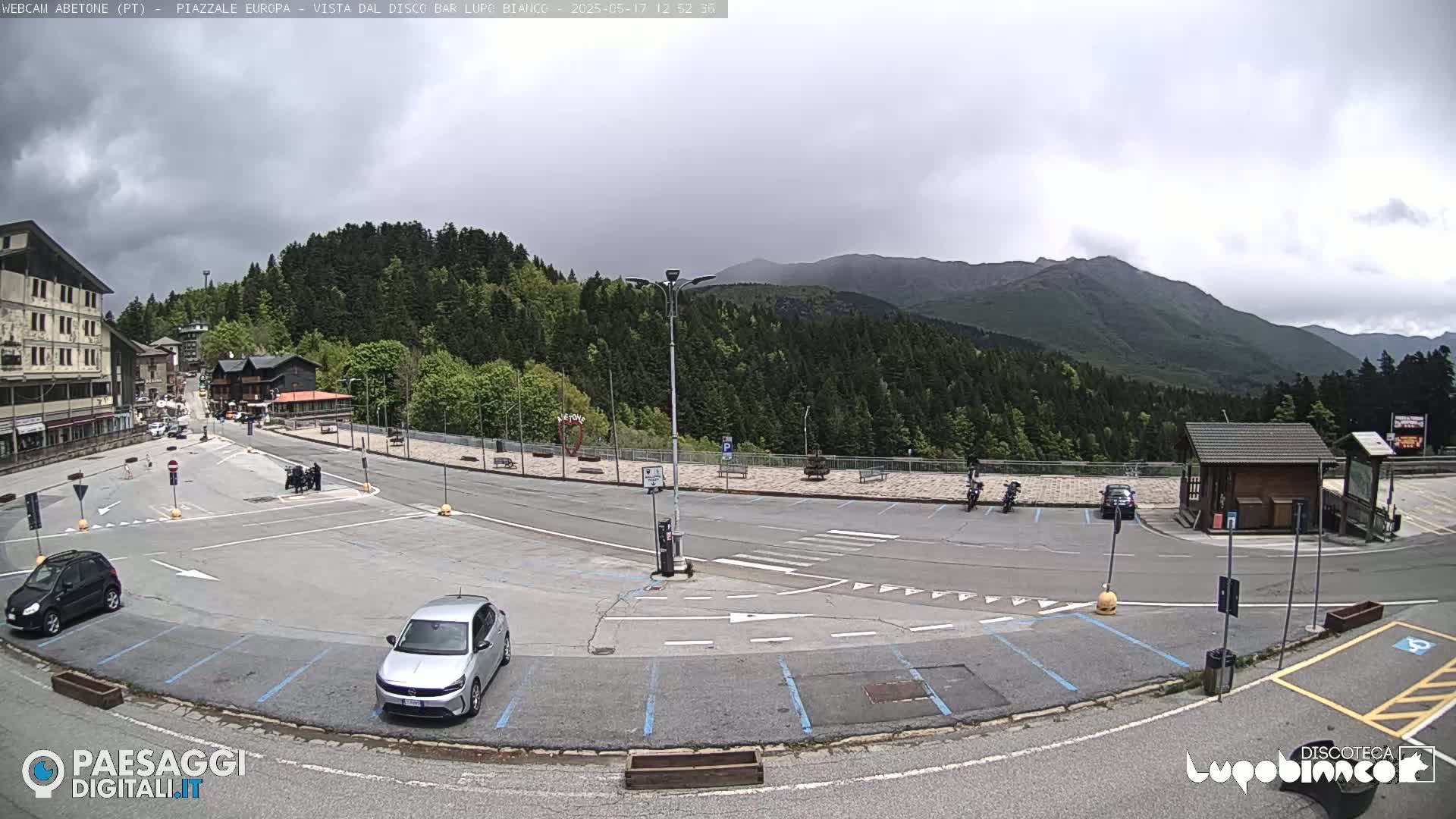 A mostly empty parking lot in a mountainous area under a cloudy sky, with a few cars and motorcycles present, and buildings in the background.