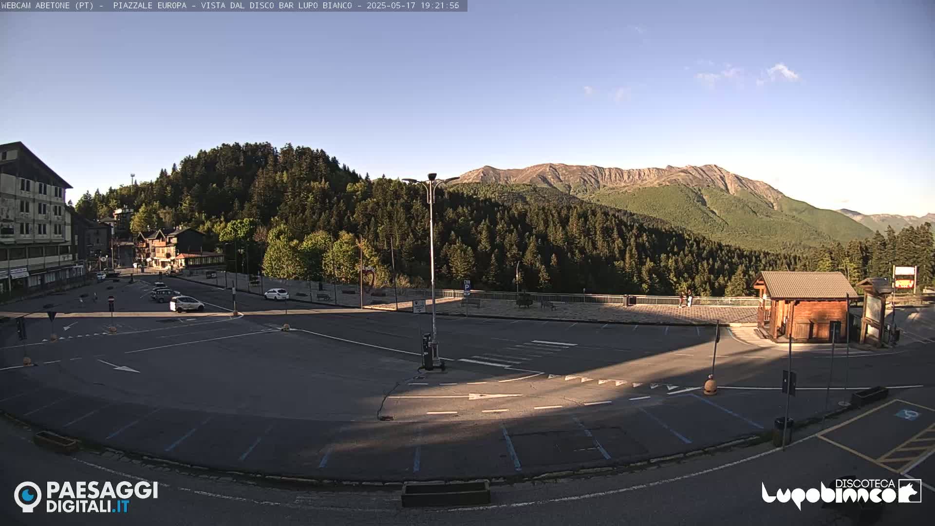 An empty parking lot sits in front of a building, with a few cars parked nearby, overlooking a mountain range under a clear, sunny sky.