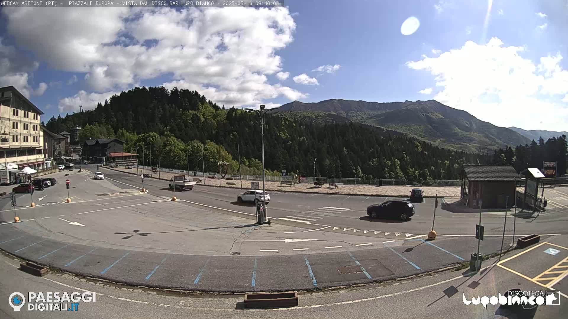 A mostly sunny day in a mountain town shows a parking lot with several cars, a roadway, and buildings, with a forest and mountains in the background.