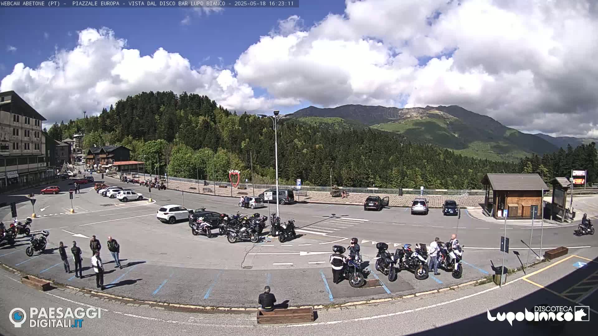 A parking lot filled with cars and motorcycles sits in front of a building and a wooded mountain range under a partly cloudy sky.
