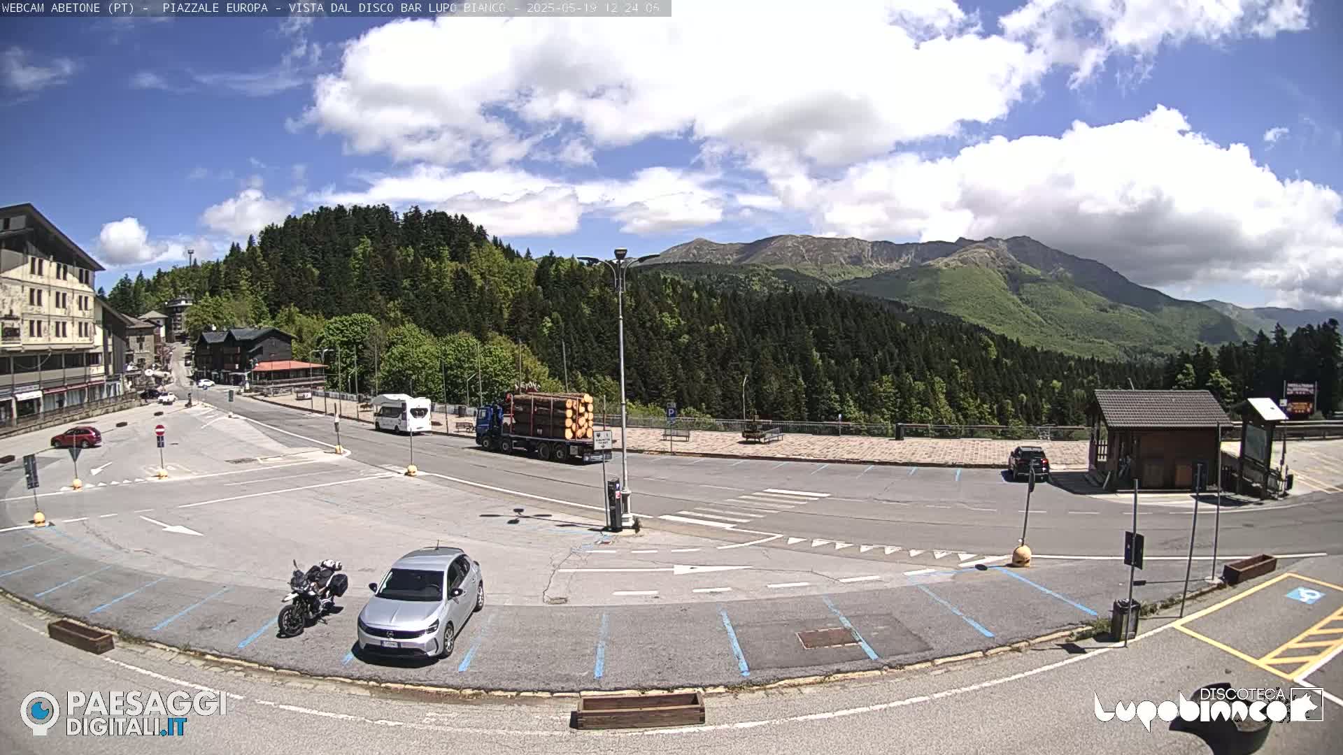 A mostly empty parking lot in a mountainous area, under a partly cloudy sky, with a few vehicles parked and a truck carrying logs.