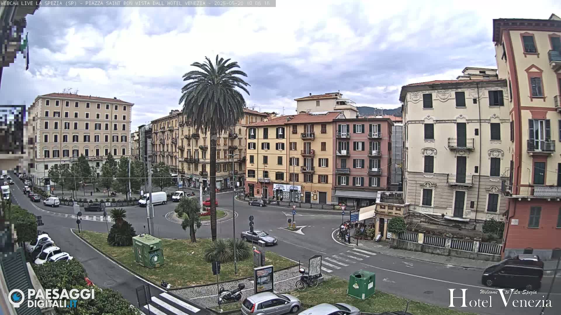 A partly cloudy day in a European city square, with several multi-story buildings surrounding a circular traffic intersection where cars are driving and people are walking.