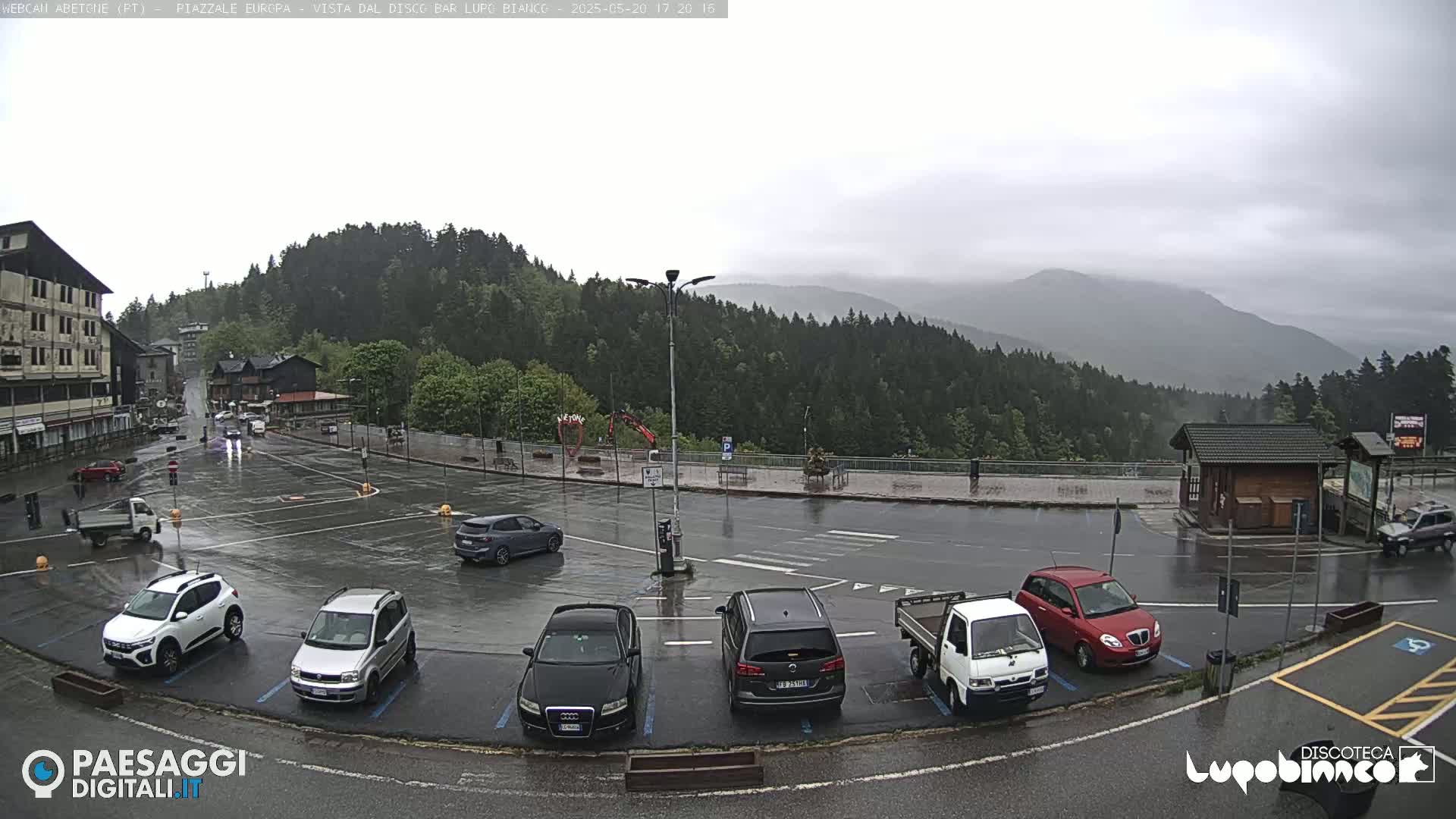 A wet parking lot with several cars is situated in front of buildings and a mountain range under an overcast sky.