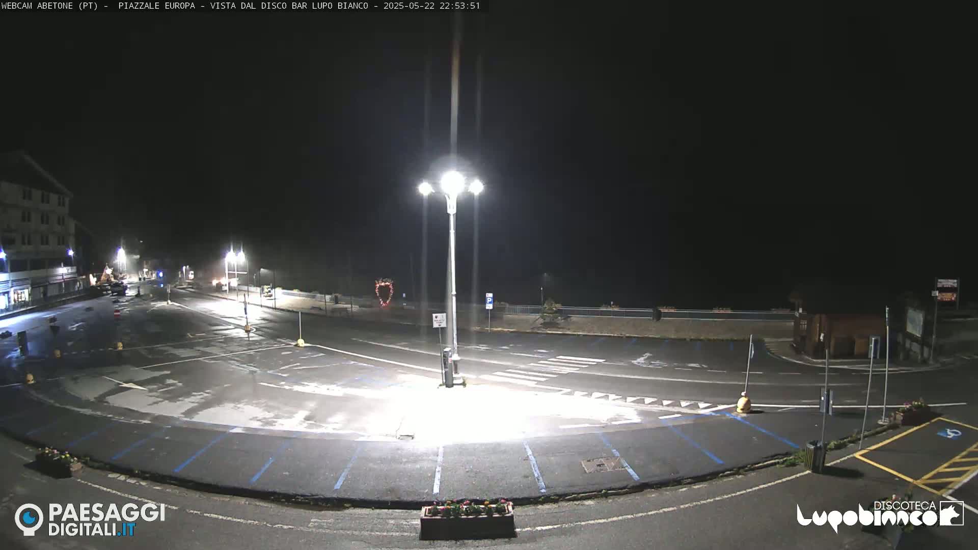 A nighttime view of an empty plaza with streetlights illuminating a mostly empty parking lot and roadways.