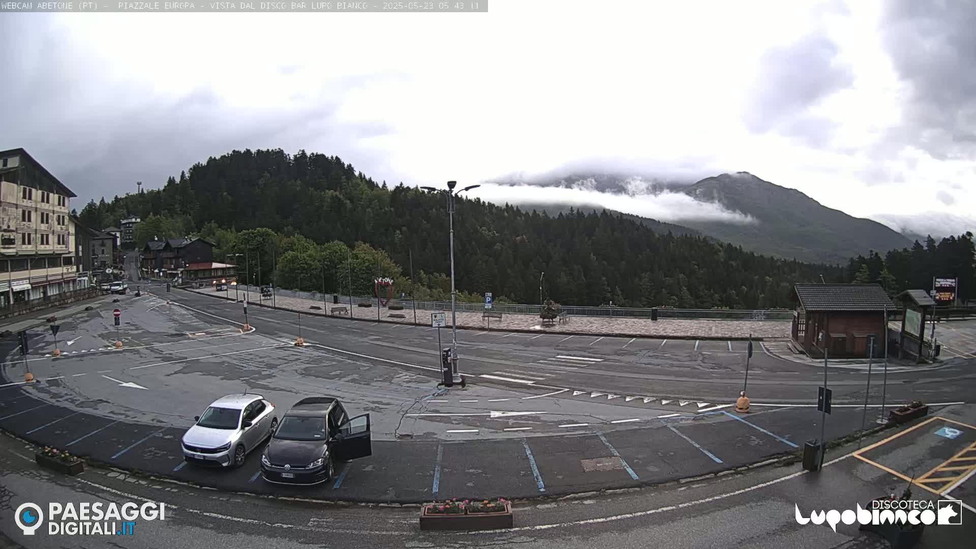 A mostly cloudy day shows a mostly empty parking lot overlooking a mountain range partially obscured by fog, with two cars parked near the lower left.