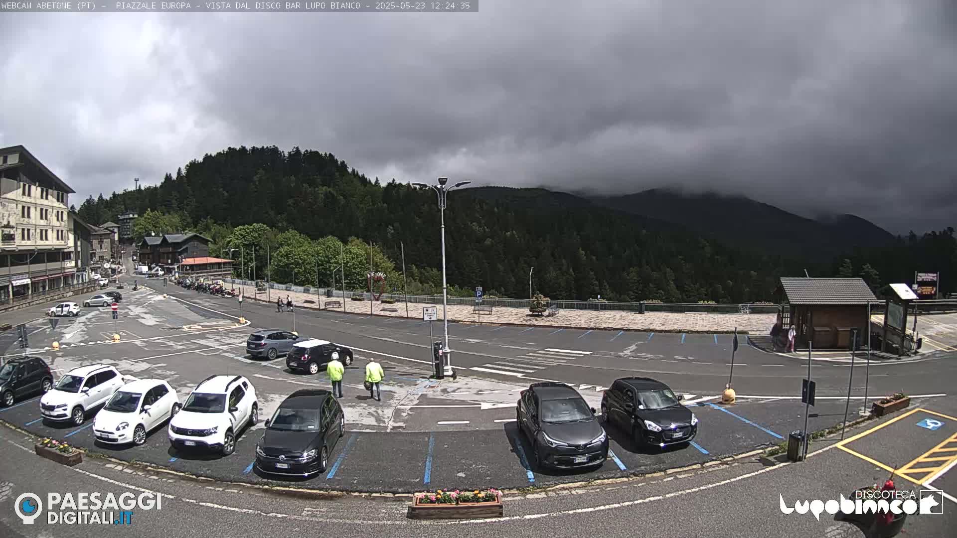A mostly cloudy day shows a parking lot filled with cars, a few people walking, and a mountainside covered in dark green trees in the background.