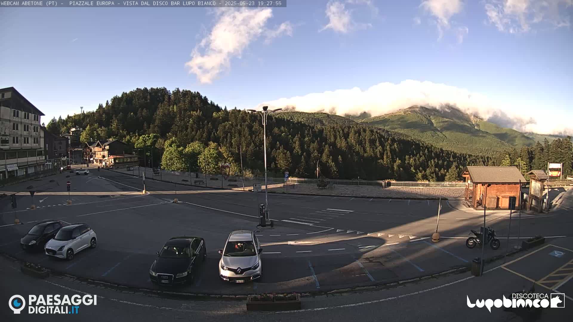 A mostly empty parking lot in a mountainous area with a few cars and a motorcycle parked, under a partly cloudy sky.