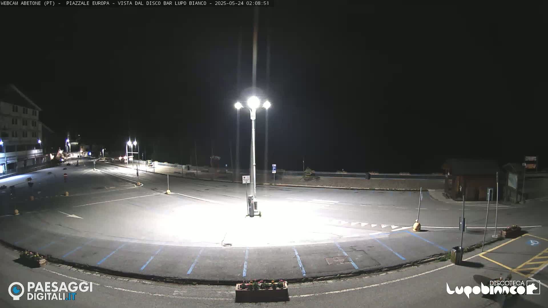 An empty, brightly lit parking lot at night is shown, next to a building and a road, under a dark, clear sky.