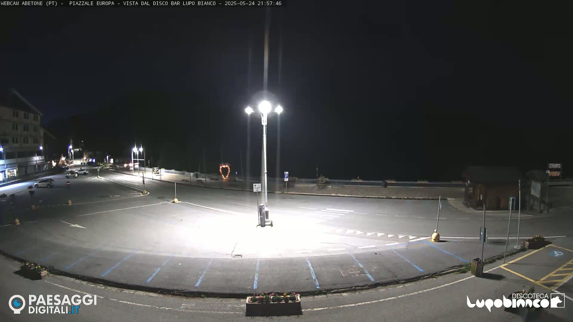 An empty, dark parking lot at night, illuminated by several streetlights, is bordered by buildings and a low wall.