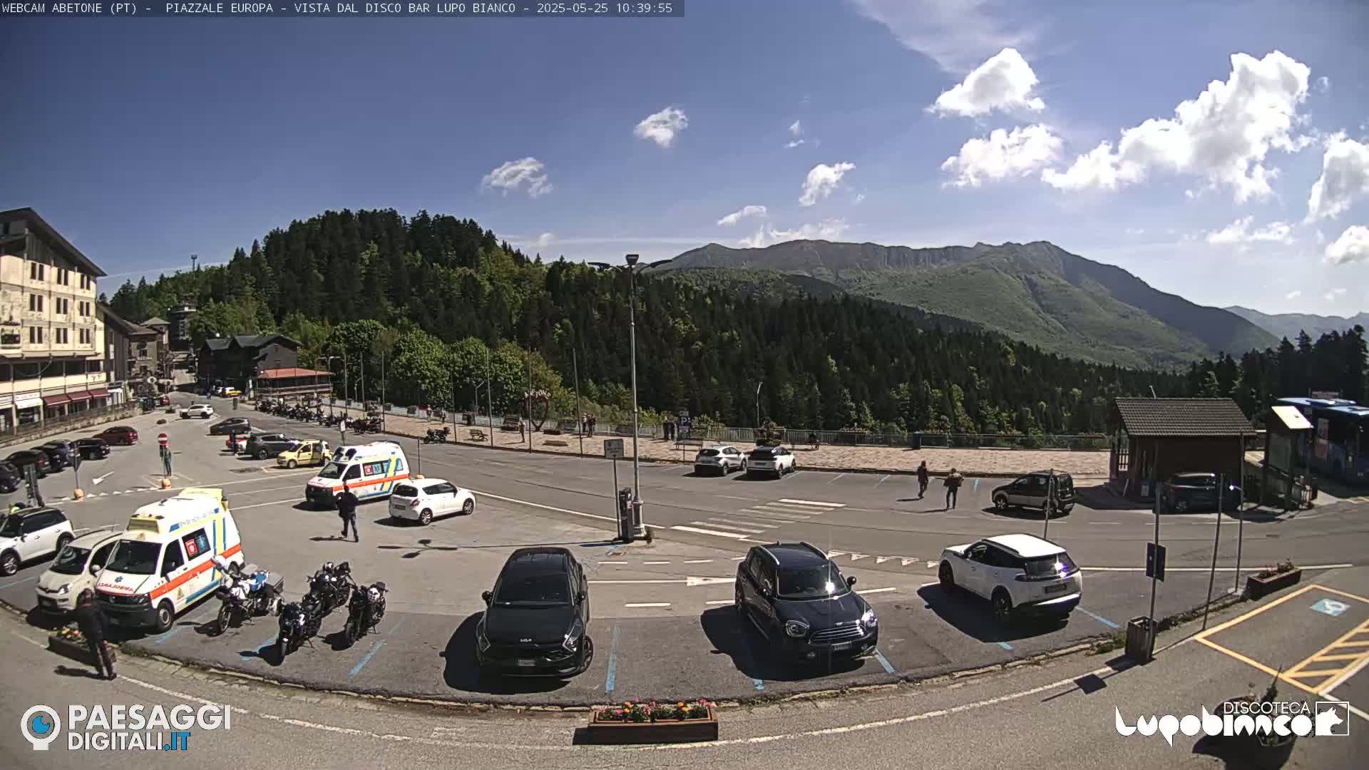 A sunny parking lot with several cars and two ambulances is situated in front of a mountain range covered in green trees and under a partly cloudy sky.