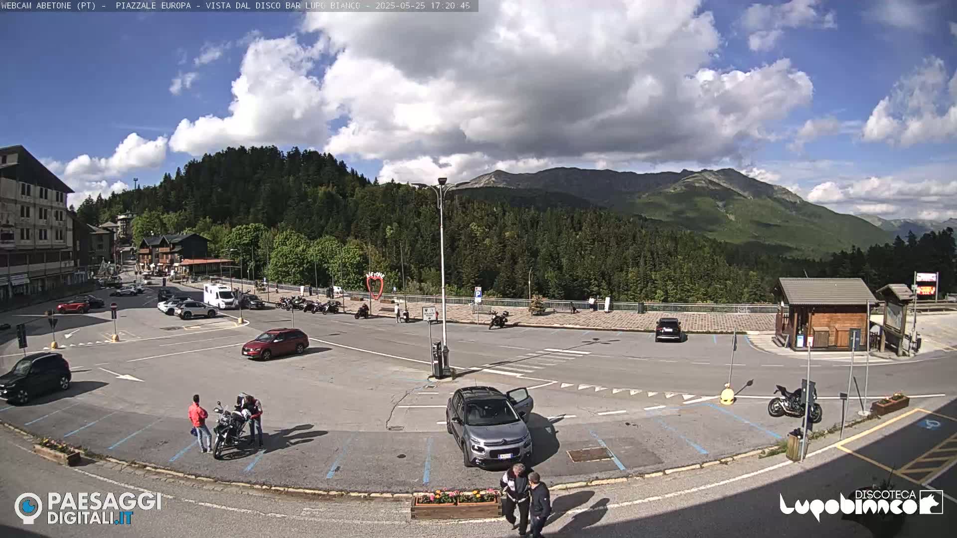 A mostly sunny day in a mountain town parking lot with several cars and motorcycles parked, and people standing near some of them; a forested mountain range is visible in the background.