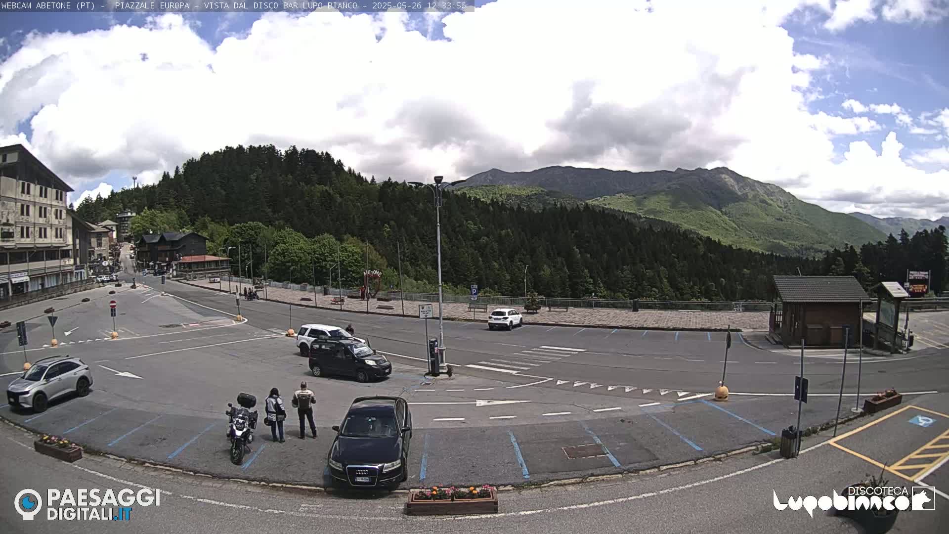 A mostly sunny day reveals a parking lot with several cars and a motorcycle, overlooked by a building and mountains in the background.