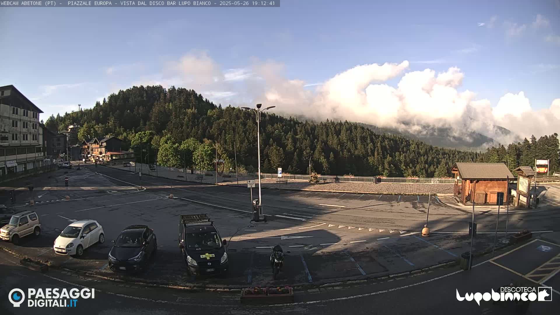 A mostly empty parking lot with several cars and a van is situated in front of a mountain range partially covered in clouds under a partly sunny sky.