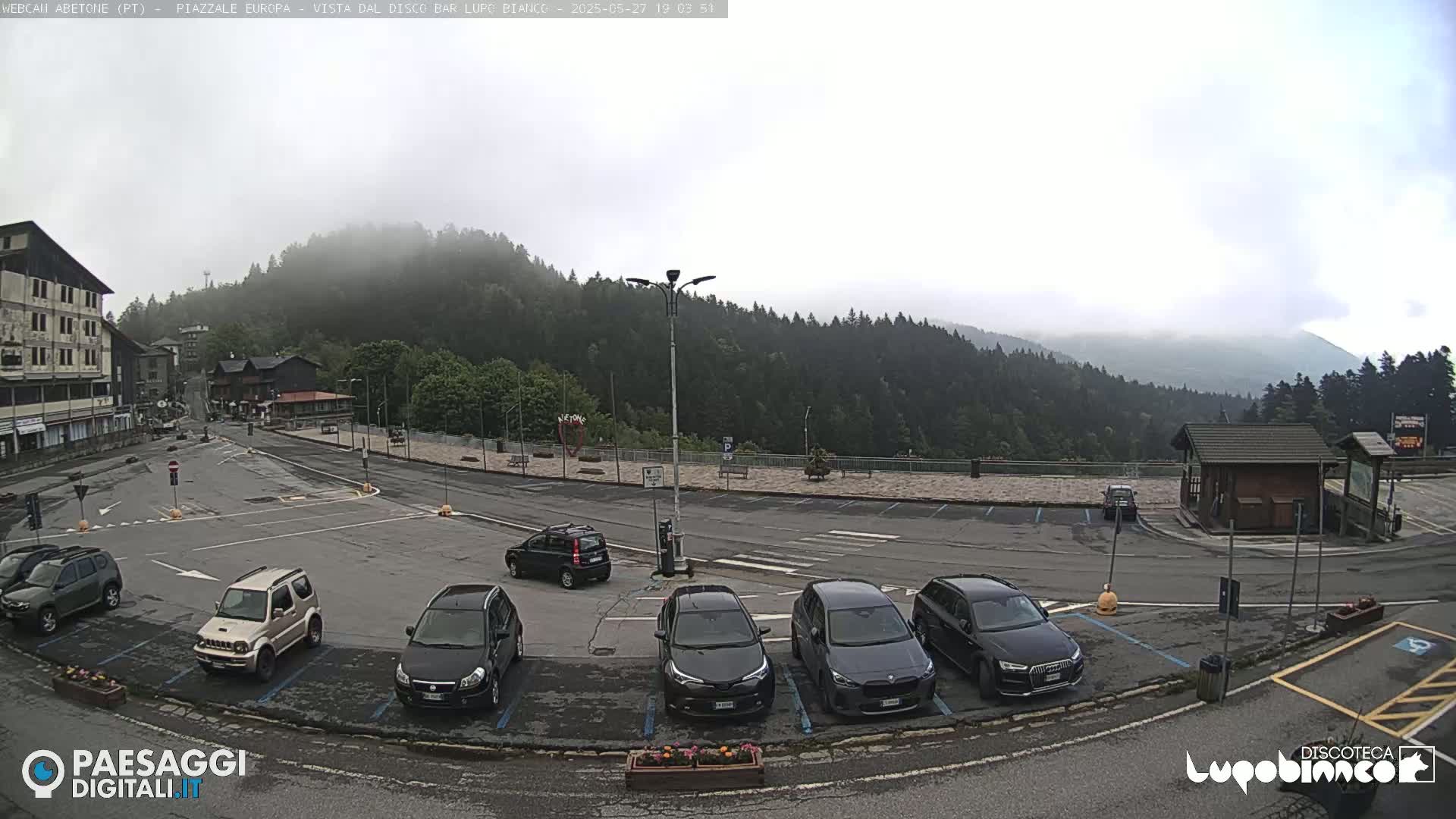 A mostly empty parking lot with several cars is situated in front of a building and overlooks a mountain range shrouded in fog.