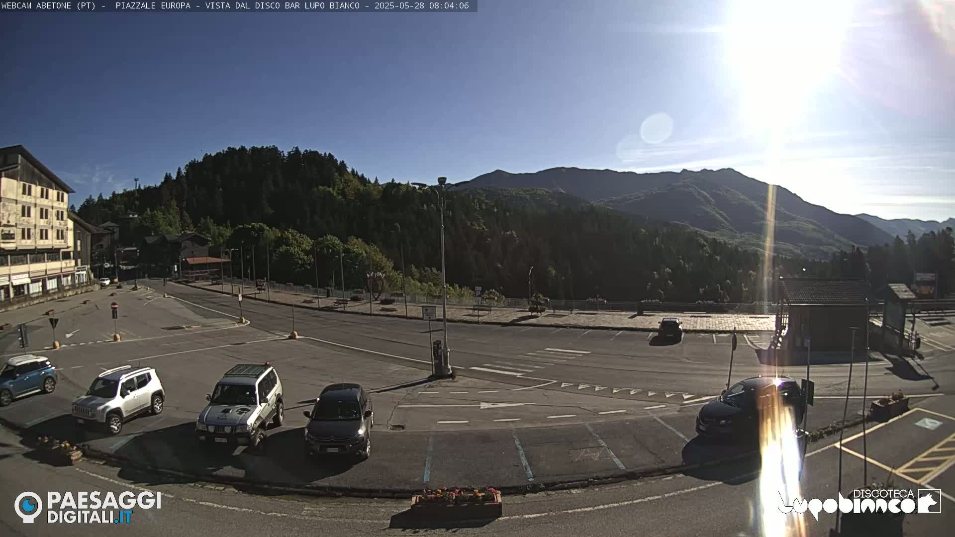 A sunny mountain vista overlooks a mostly empty parking lot with several cars, a building, and some greenery.