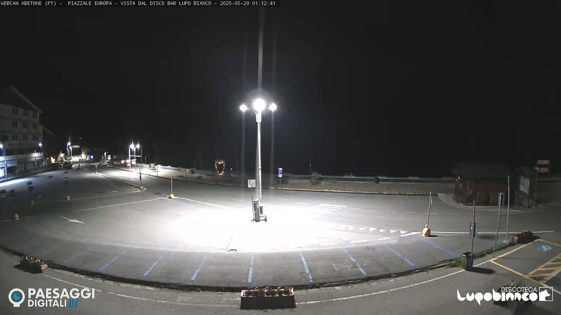 An empty, brightly lit parking lot at night is shown, with a building and a few cars visible in the distance.