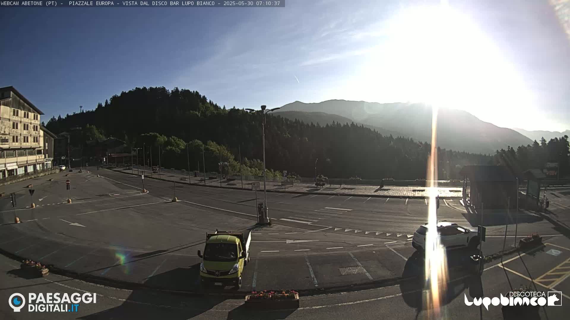 A mostly empty parking lot sits in front of a building, overlooking a mountain range under a mostly clear, sunny sky.