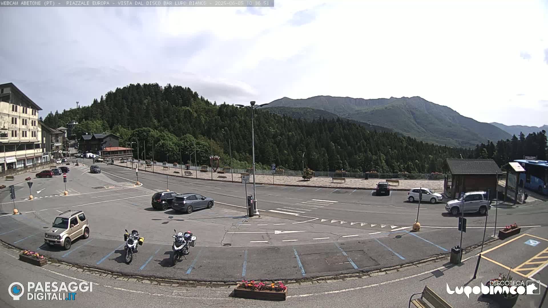 A mostly empty parking lot with several cars and two motorcycles is situated in front of a mountain range under a partly cloudy sky.