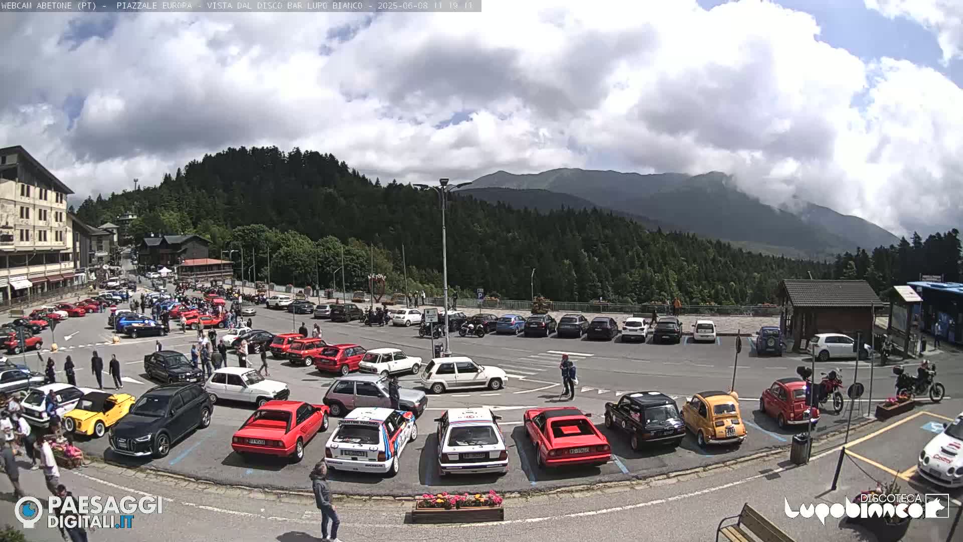 A parking lot filled with various cars is situated in a mountainous area under a partly cloudy sky.