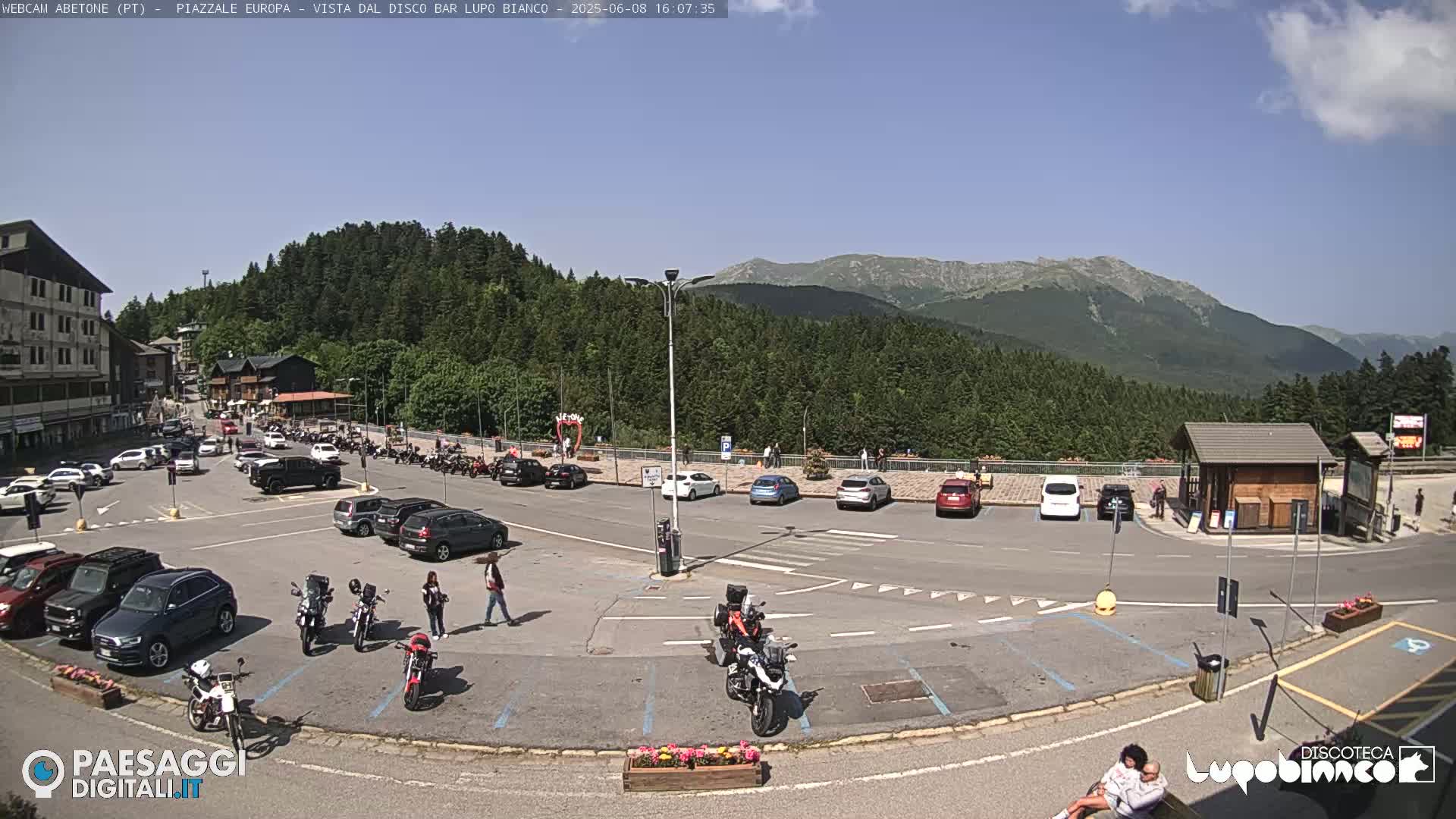 A sunny mountain town parking lot with numerous cars and motorcycles parked, surrounded by evergreen trees and mountains in the background.