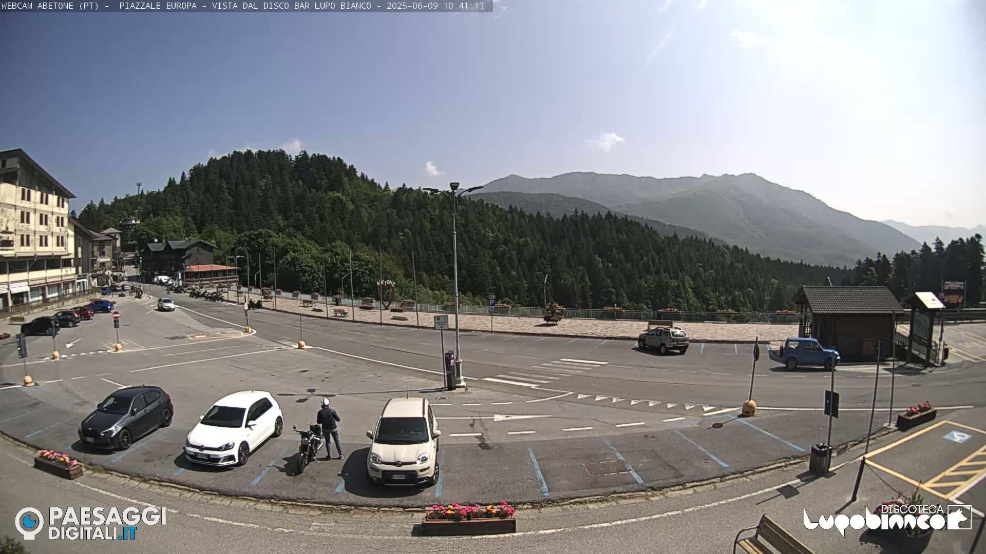 A mostly empty parking lot is situated in a mountain town, with cars parked and a few people visible, under a clear, sunny sky and a backdrop of green, forested mountains.