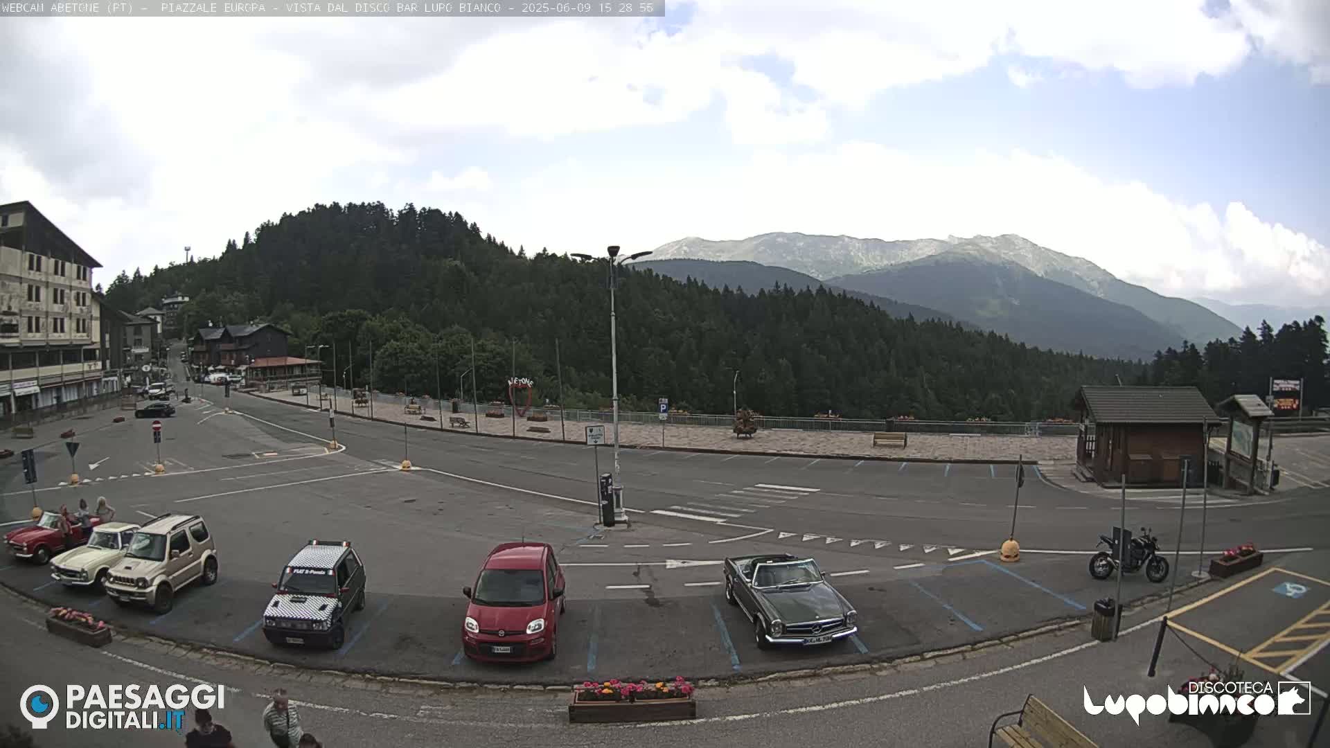 A mostly empty parking lot with several cars and a motorcycle is situated in front of buildings and a forested mountain range under a partly cloudy sky.