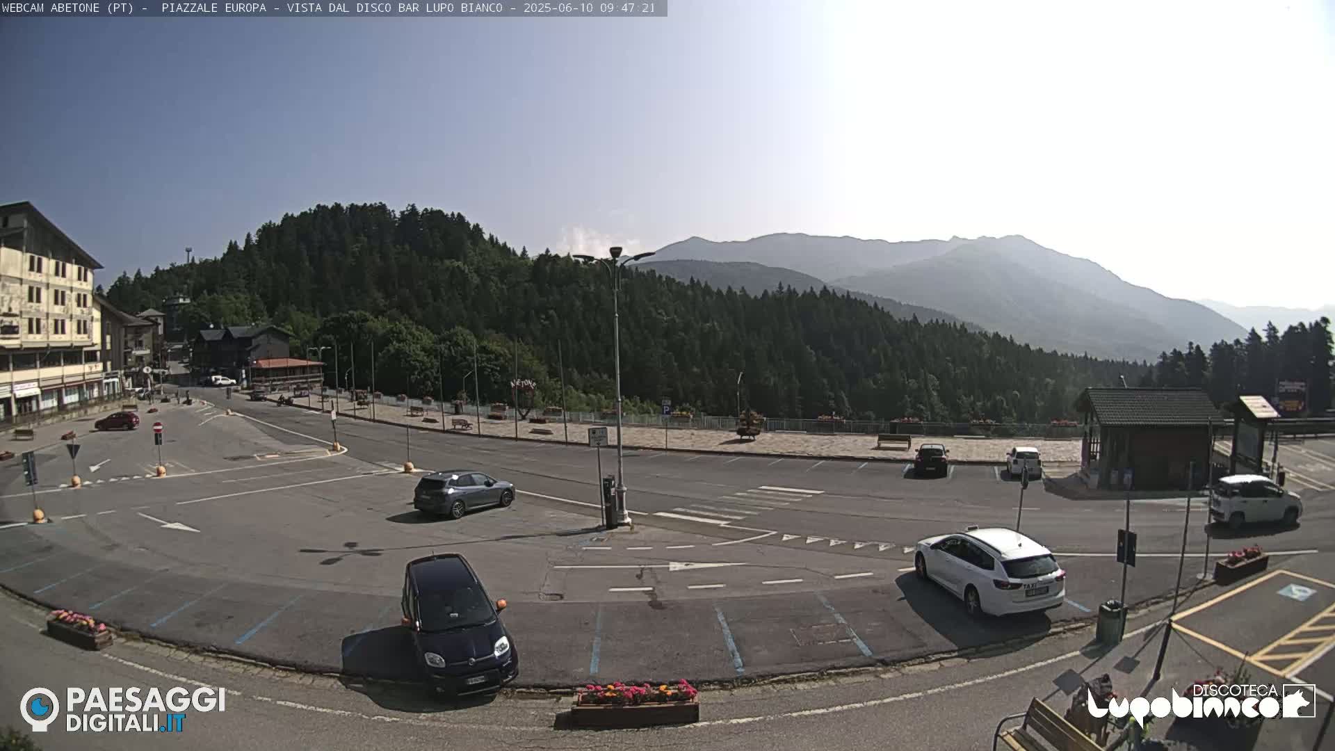 A mostly empty parking lot with several cars is situated in front of a building, with a mountainous landscape and a largely clear sky in the background.