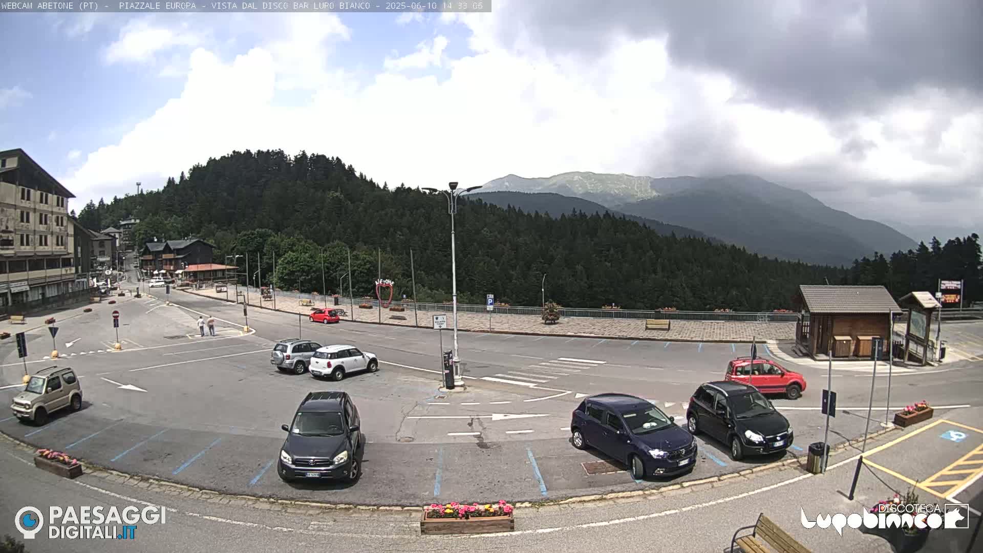 A mostly cloudy day reveals a mountain vista overlooking a parking lot with several cars, a few pedestrians, and small buildings.