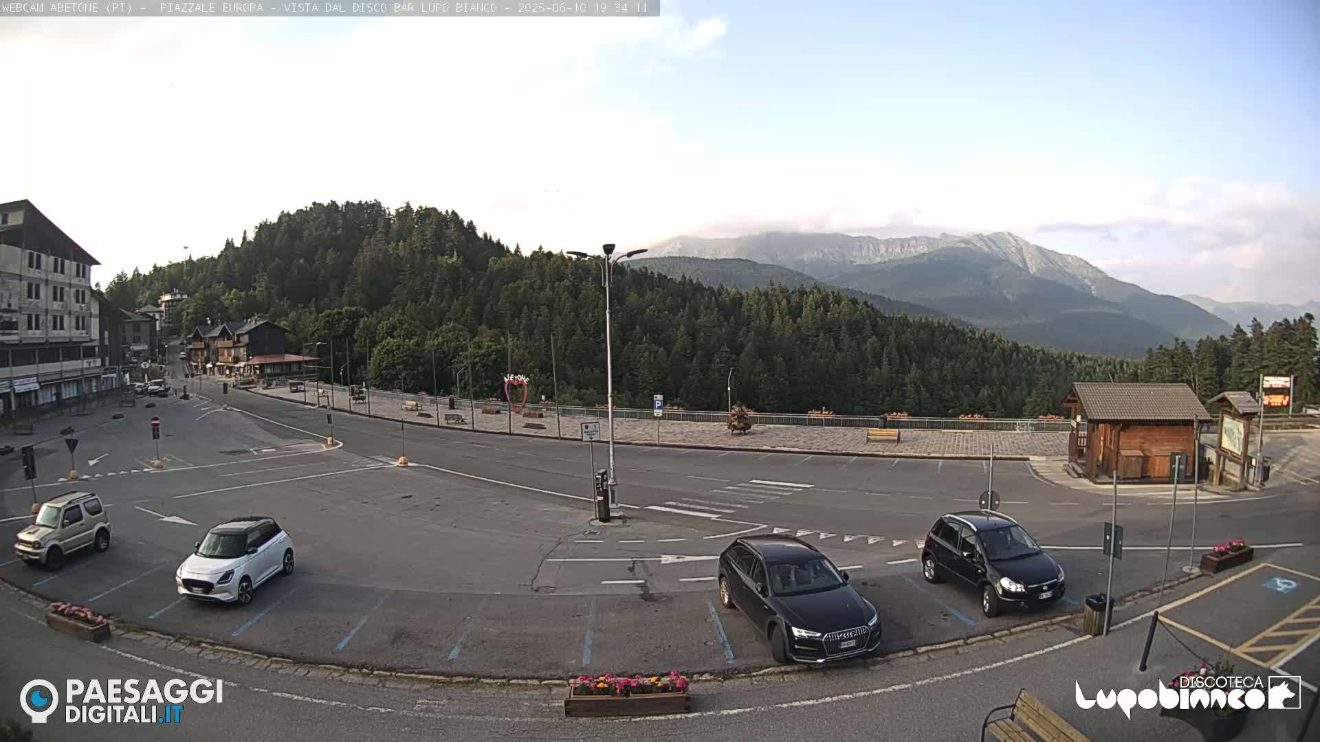 A mostly empty parking lot in a mountainous area with several cars parked, overlooked by a building and a wooden kiosk under a partly cloudy sky.
