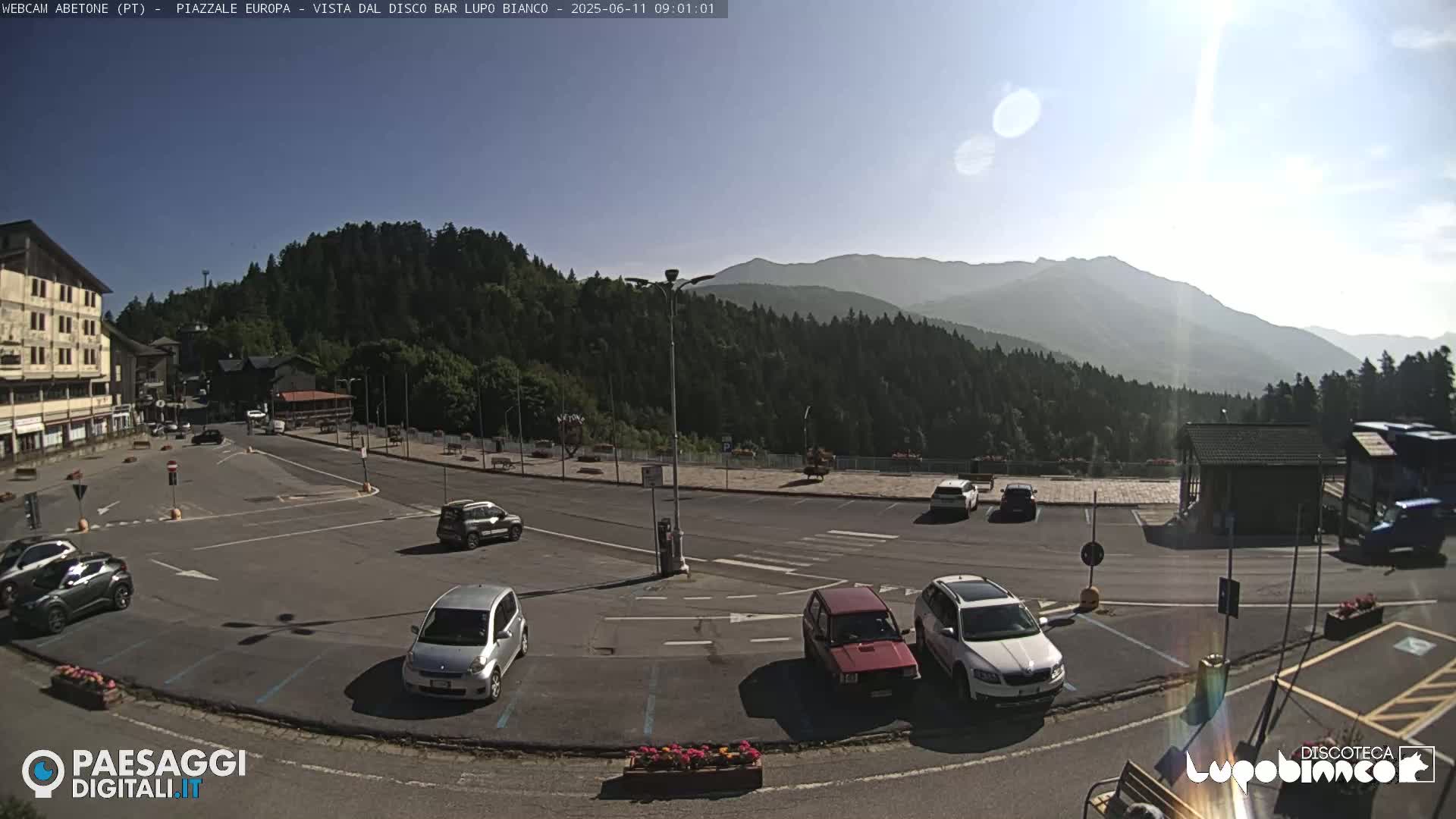A sunny mountainside parking lot with several cars, a few buildings, and a large forested mountain in the background.