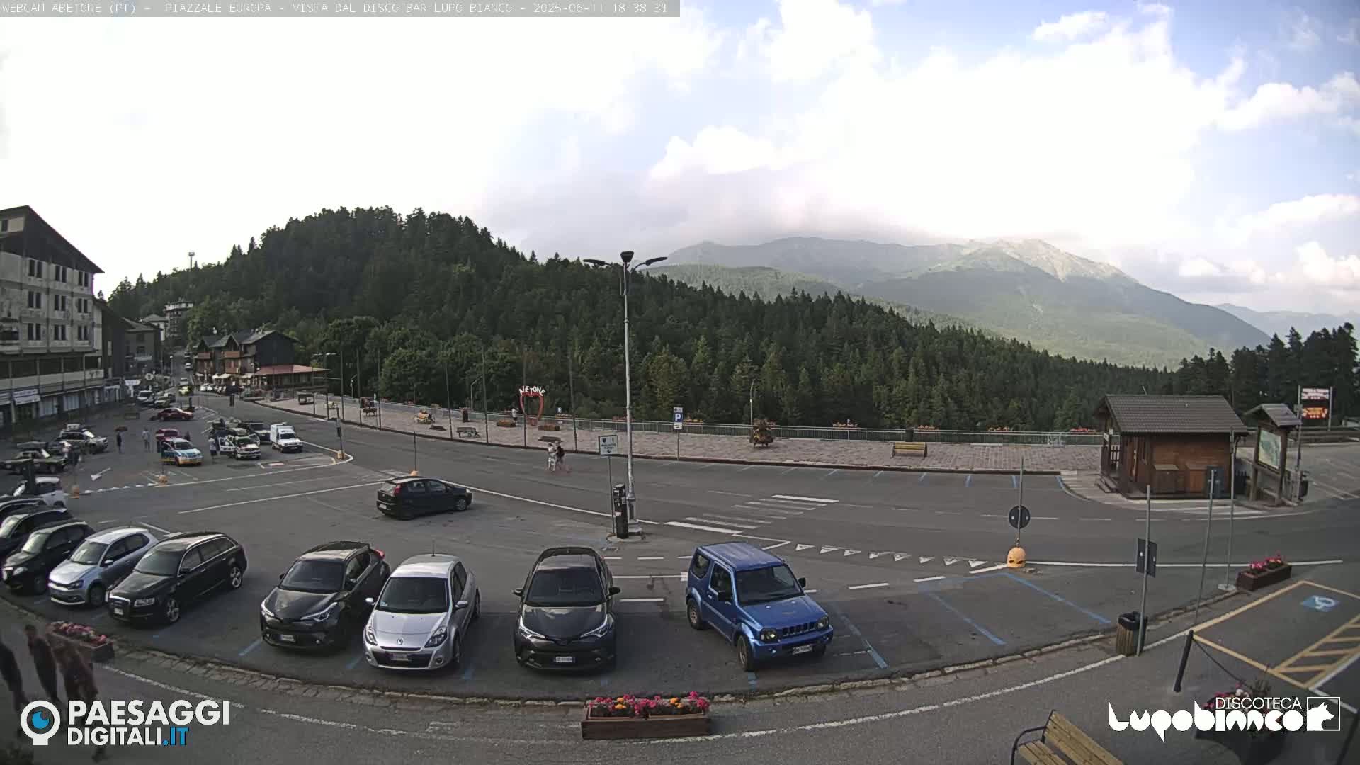 A mostly empty parking lot with several cars is situated in front of a building and a wooded mountain range under a partly cloudy sky.