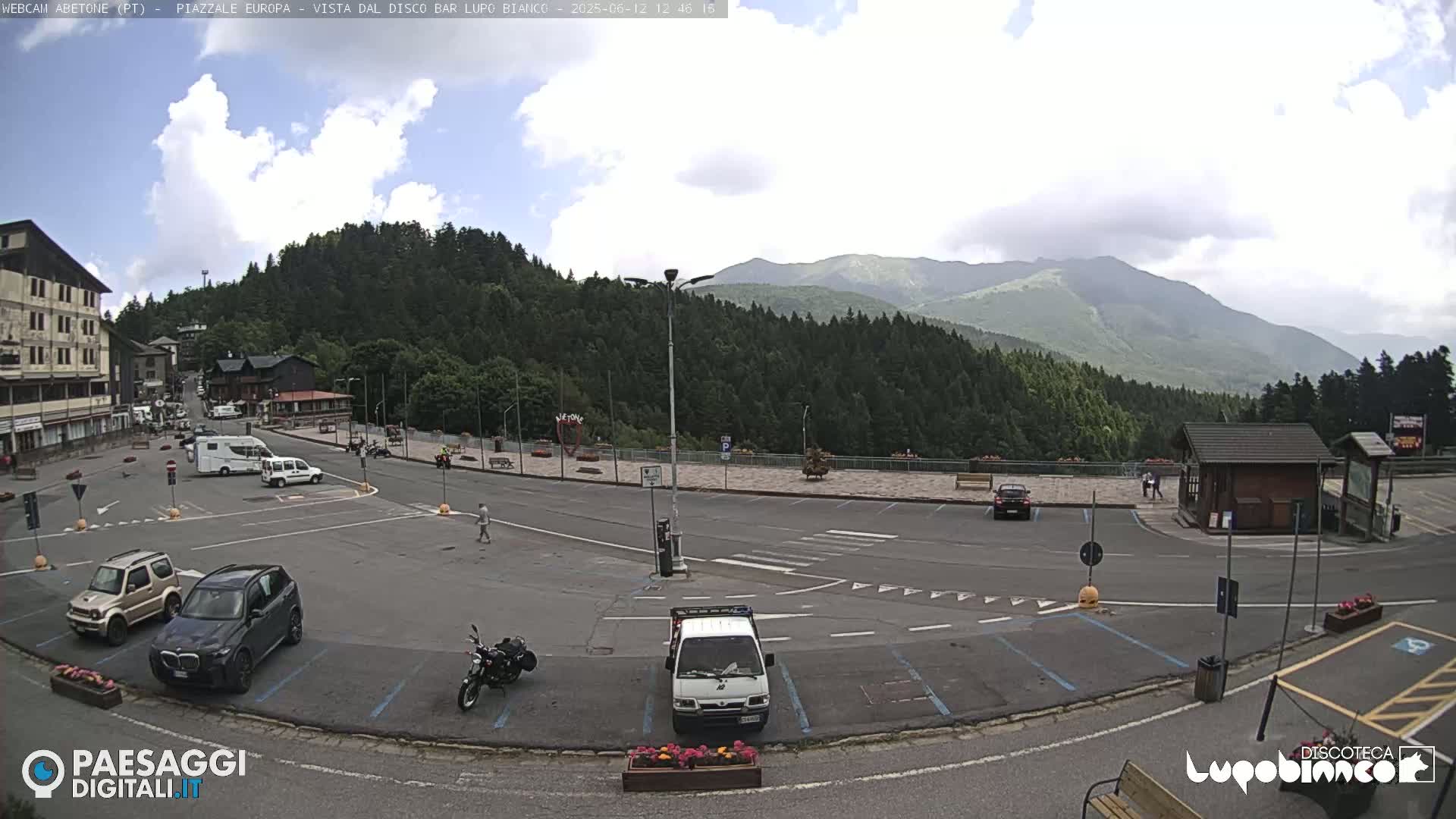 A partly cloudy day reveals a mountain view overlooking a parking lot with several vehicles, a few pedestrians, and a small building.