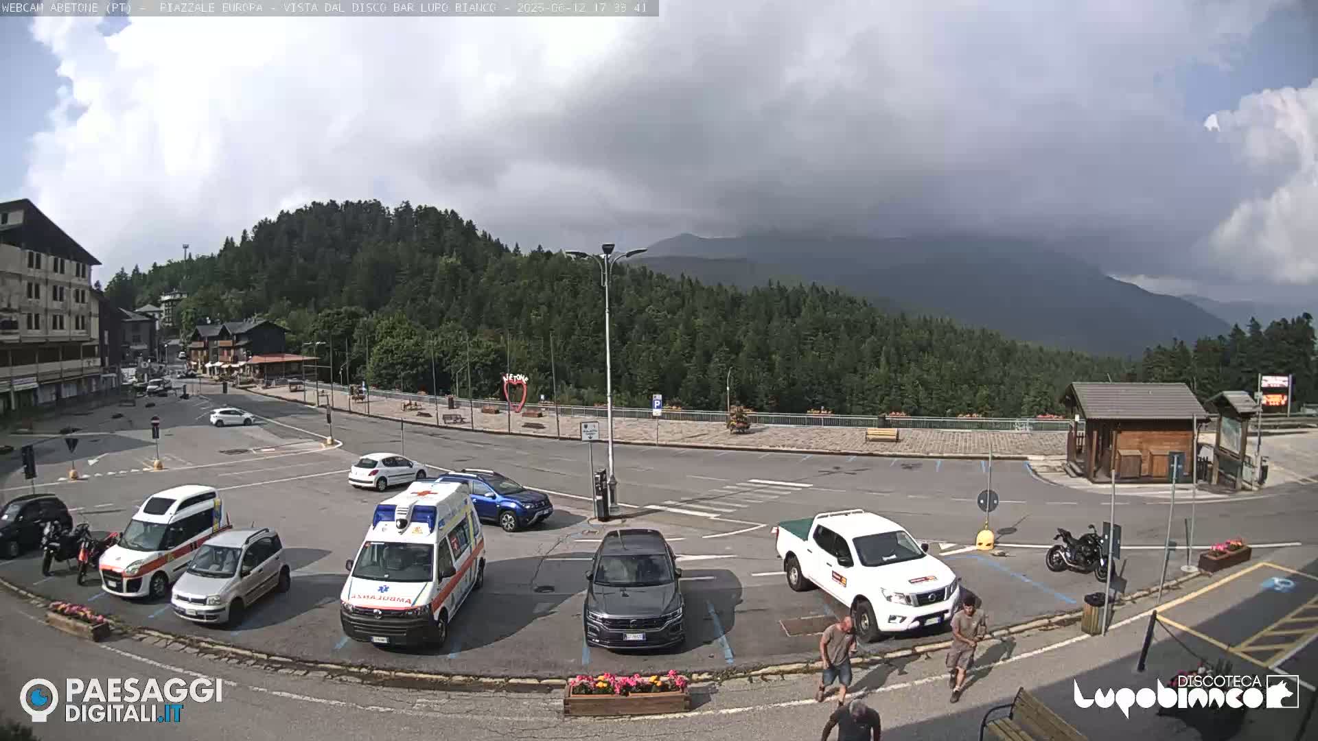 A mostly empty parking lot with several vehicles, including an ambulance, is situated in a mountain town under a cloudy sky.