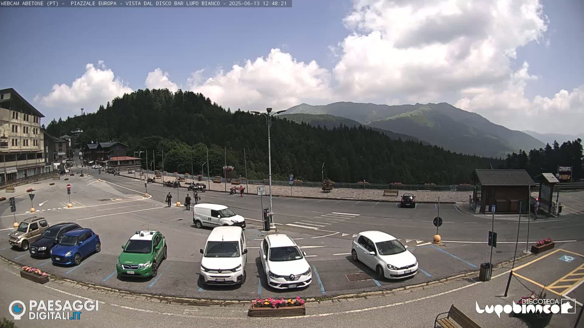 A mostly sunny parking lot with several cars and vans is situated in a mountain town, with a mountain range and forest in the background.